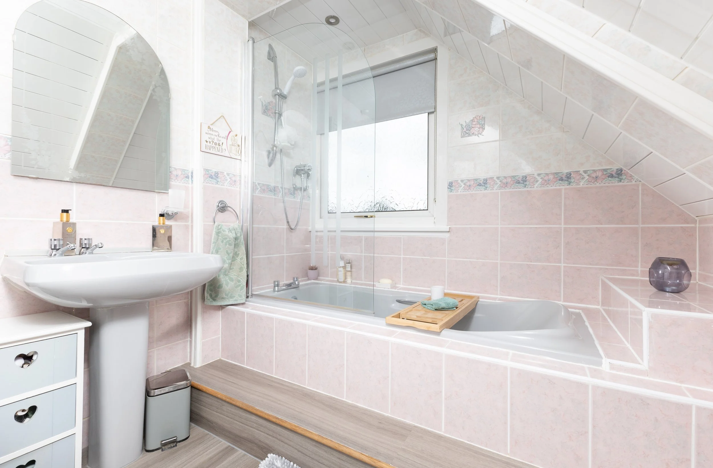 A bathroom with a bathtub, glass shower door, sink, mirror, and various toiletries. The bathroom has pink tiled walls with a floral border, a window with a roller blind, and a sloped ceiling.