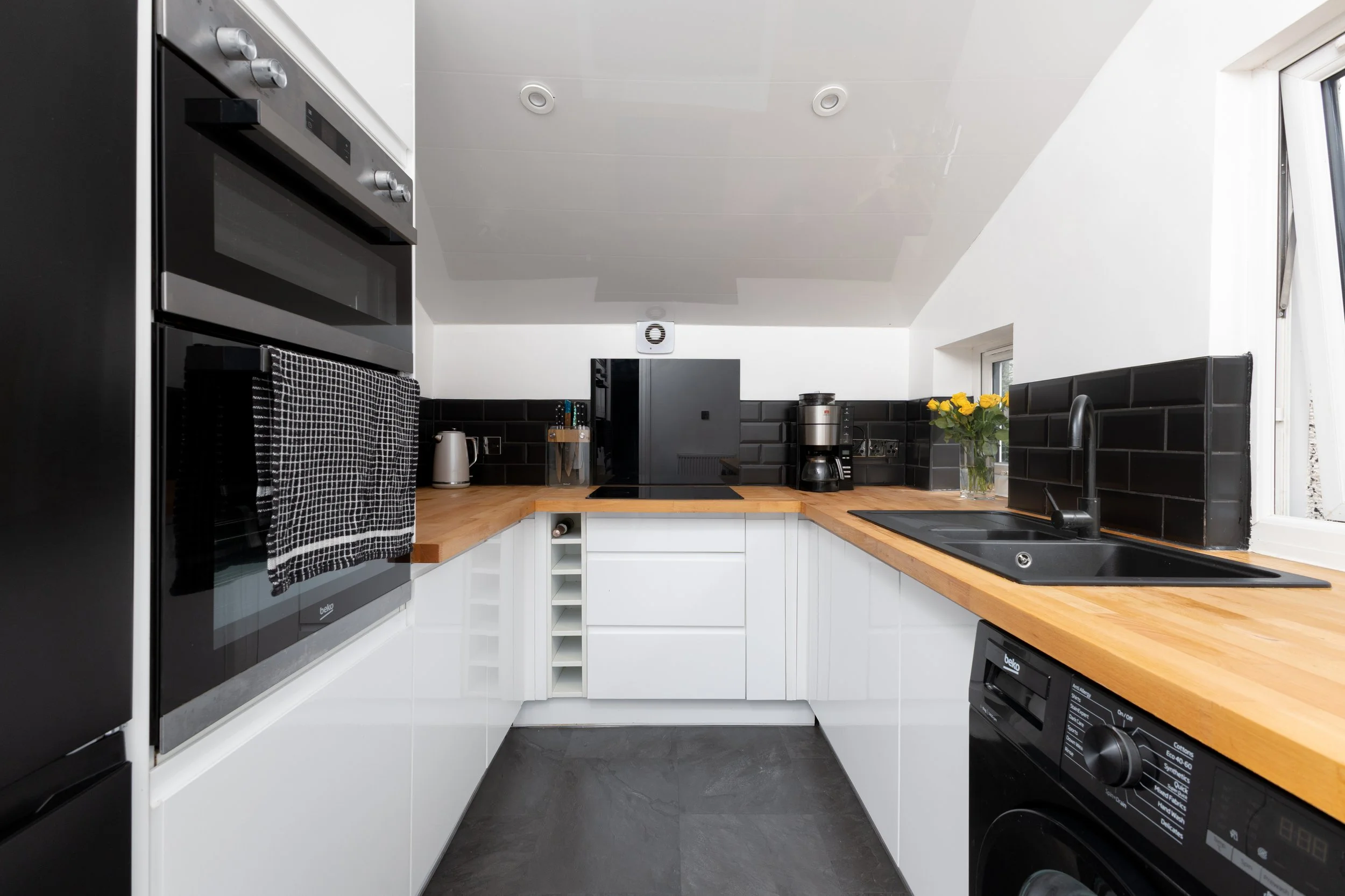 Modern kitchen with white cabinets, black backsplash, and wooden countertops, featuring appliances like an oven, coffee maker, and washing machine.