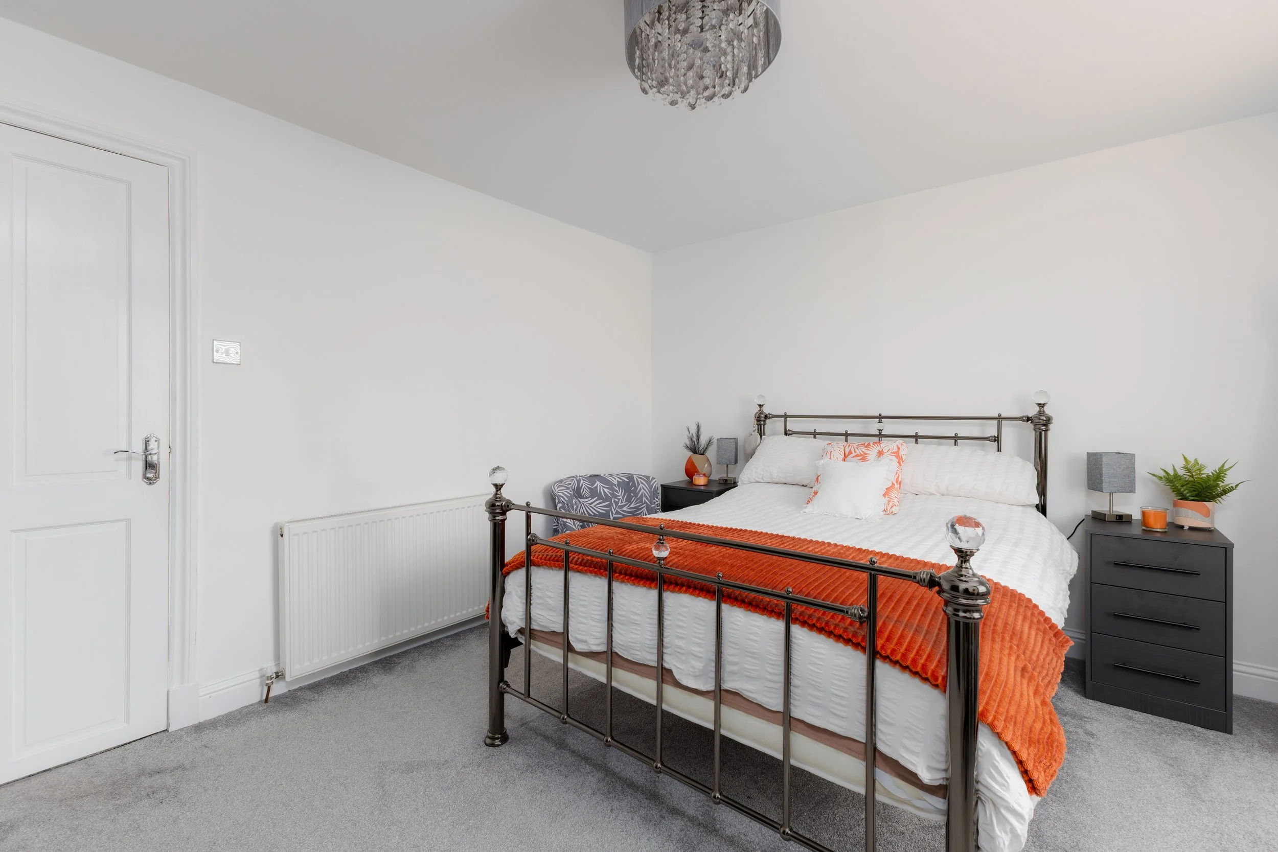 A bedroom with white walls, a metal bed frame with glass finials, white bedding, orange throw blanket, and pillows. Two gray nightstands with lamps, a small plant, and a decorative item are on each side of the bed. A ceiling light fixture and a white