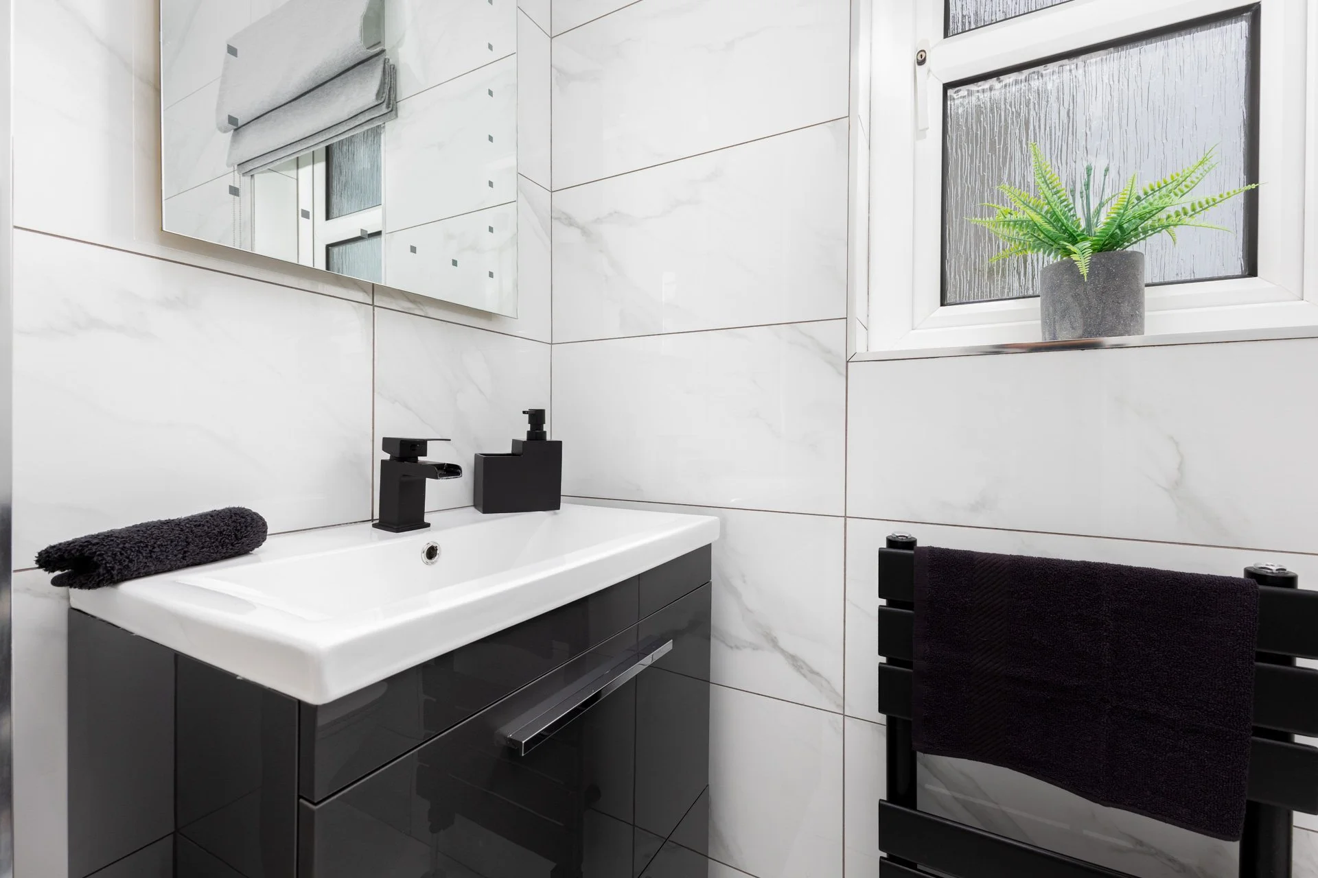 Modern bathroom with white marble tiles, black sink vanity, black faucet, a mirror, a small potted fern on the window sill, and black towels hanging on a black towel rack.