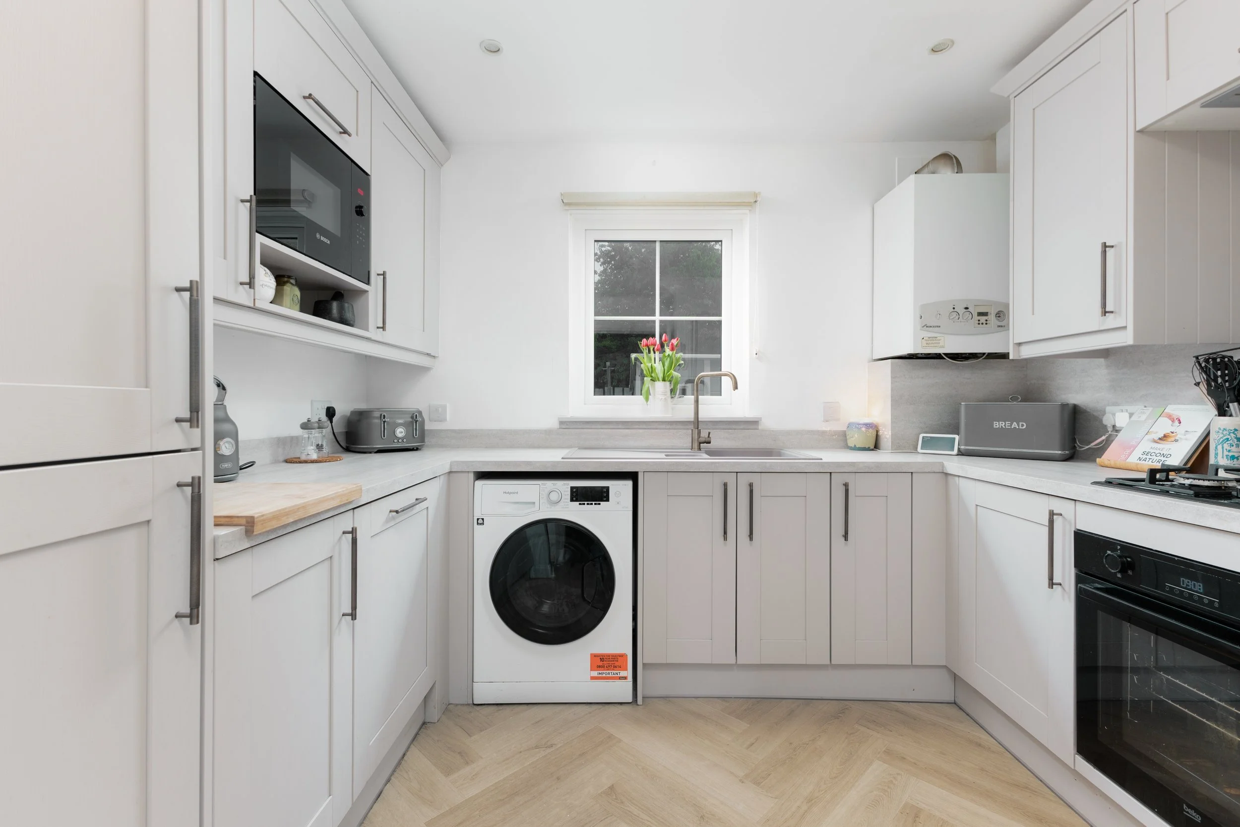 Modern kitchen with white cabinets, a window with a vase of tulips, appliances like a microwave, toaster, washing machine, gas stove, and countertop with a loaf of bread and a book.
