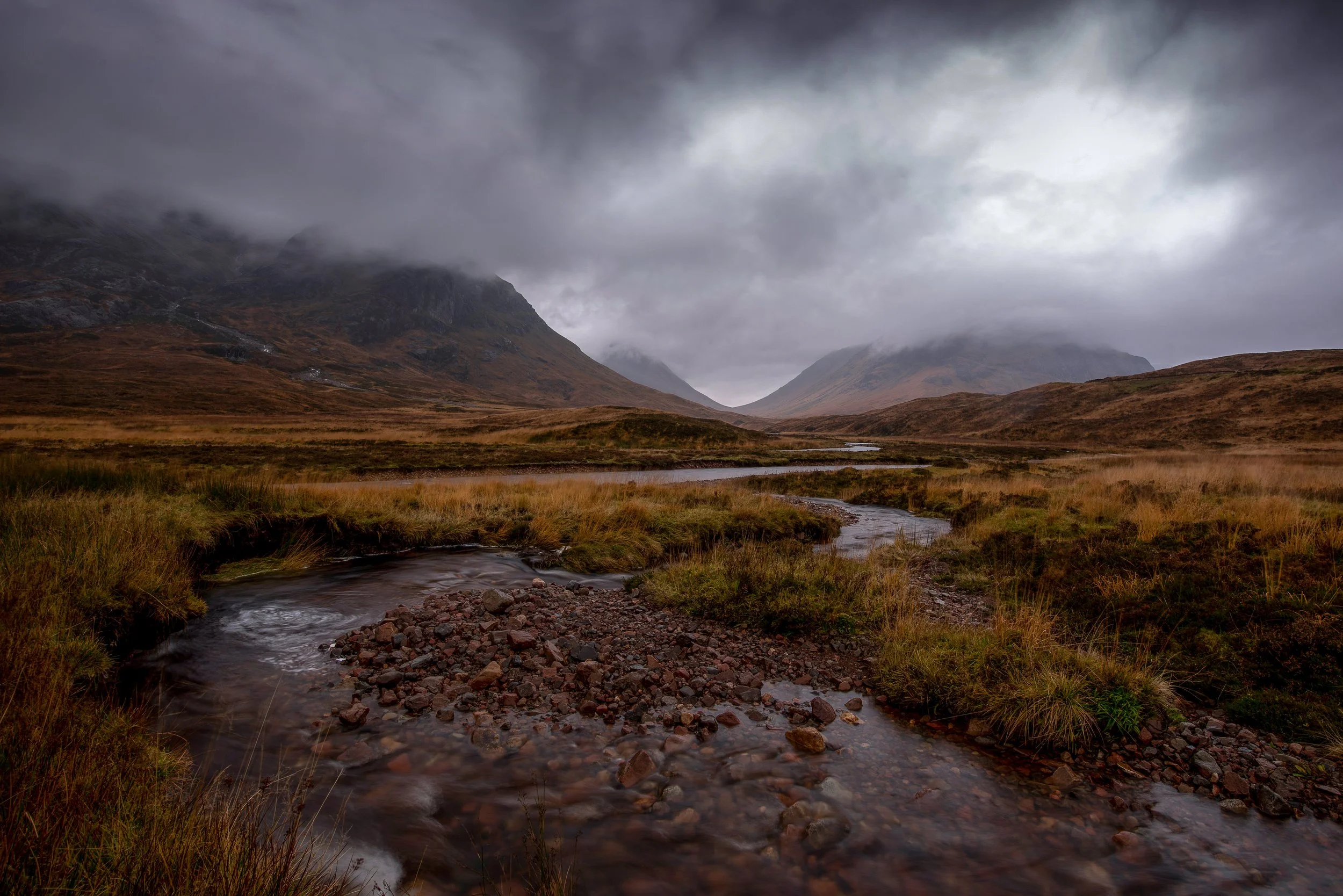 A mountain valley with dark, cloudy skies, a meandering stream flowing through grassy terrain, surrounded by rugged hills and mountains shrouded in fog.
