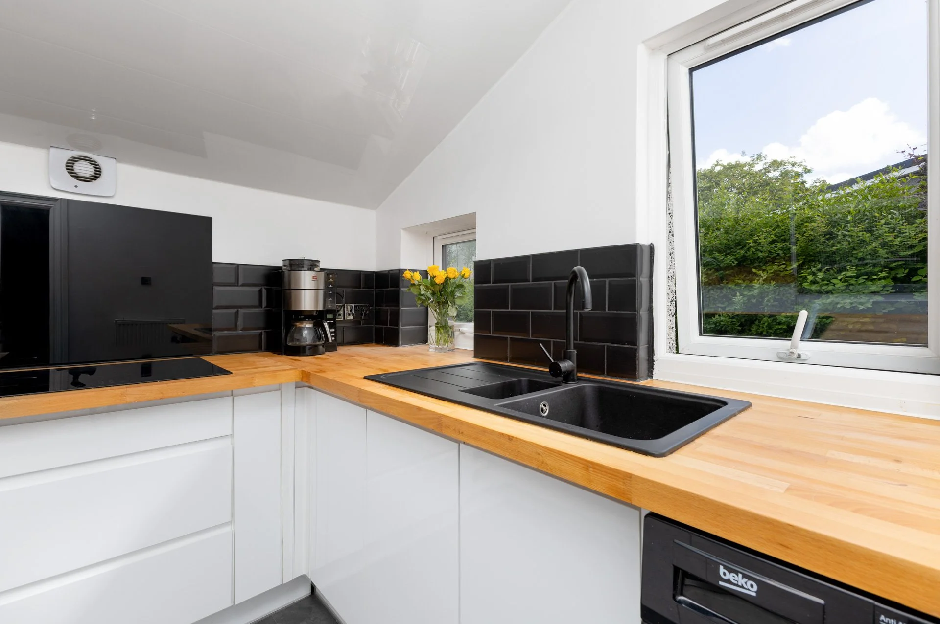 Kitchen with white cabinets, black tile backsplash, a wooden countertop, a black sink, a coffee maker, yellow flowers, large windows showing greenery outdoors, and a black dishwasher.