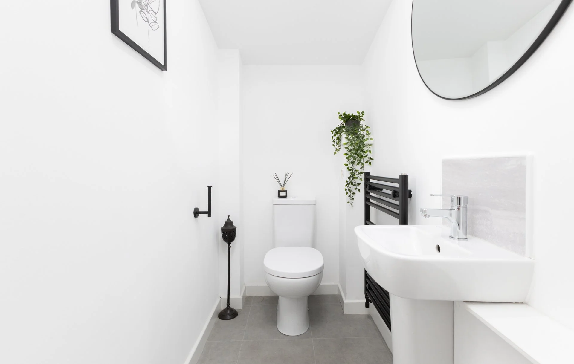Minimalist white bathroom with toilet, sink, black towel rack, mirror, potted plant, and decor, with gray floor tiles.