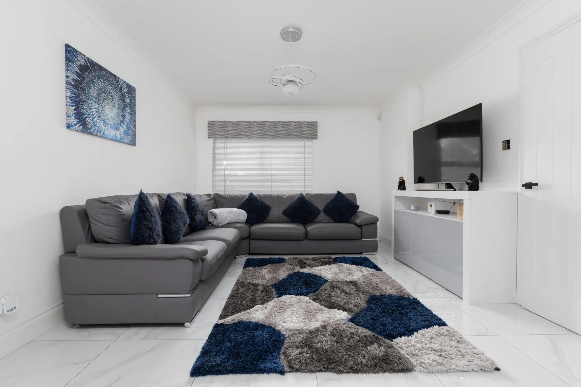 Living room with a gray sectional sofa, blue and gray throw pillows, abstract area rug, white media console, wall-mounted TV, window with blinds and valance, modern ceiling light fixture.