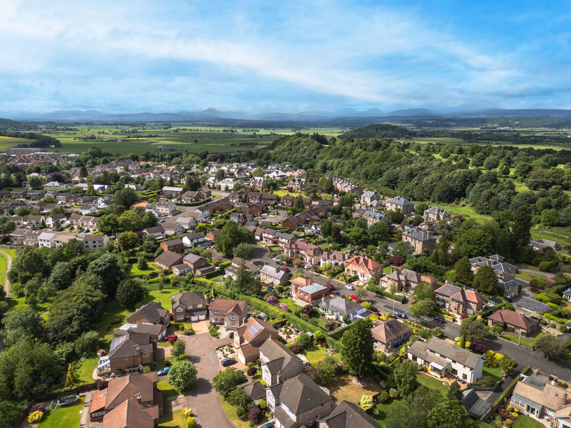 Aerial view of a suburban neighborhood with houses, lawns, and trees, surrounded by green fields and distant mountains under a partly cloudy sky.