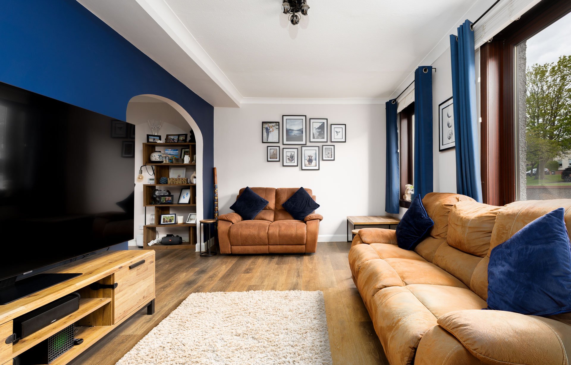 Living room with two tan sofas with blue pillows, a white shaggy rug, large windows with blue curtains, framed pictures on white walls, a wooden entertainment stand, TV, and a ceiling light fixture.