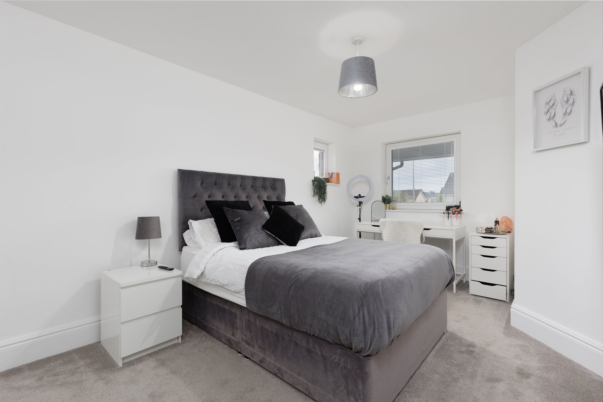 A modern bedroom with a large gray upholstered bed, white nightstand, and decor including a desk with a ring light, vanity, and artwork on white walls.
