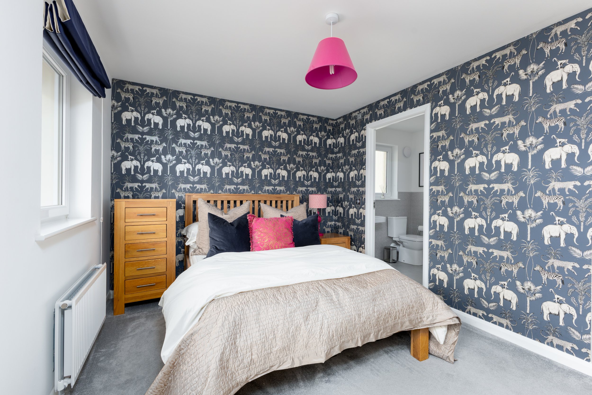 A bedroom with a wooden bed, bedding, decorative pillows, and a navy and white animal print wallpaper featuring elephants, zebras, and palm trees. There is a pink lampshade on the ceiling and a small bathroom with a toilet visible through an open doo