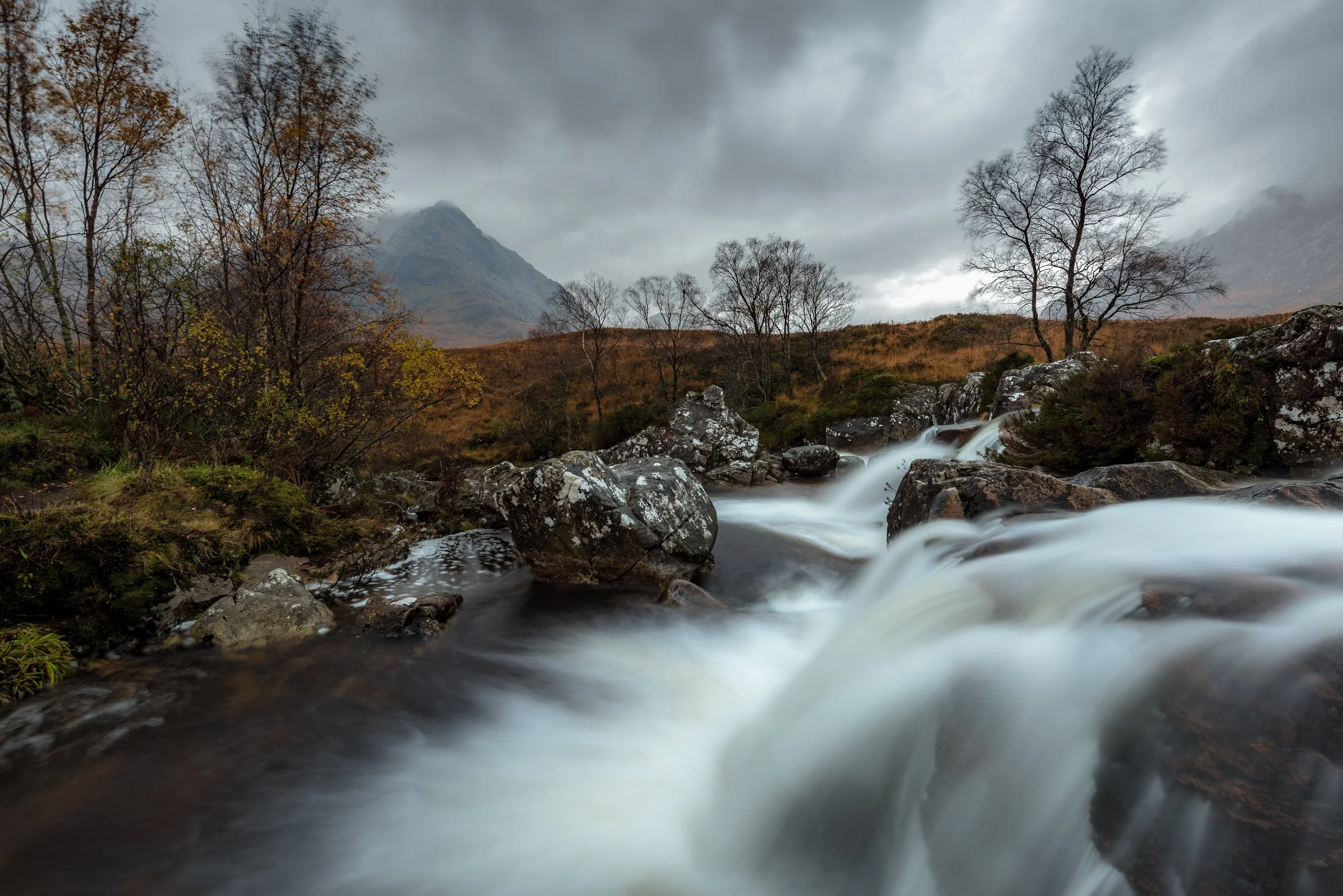 A mountainous landscape with a cloudy sky, leafless trees, and a flowing stream with rocks in the foreground.