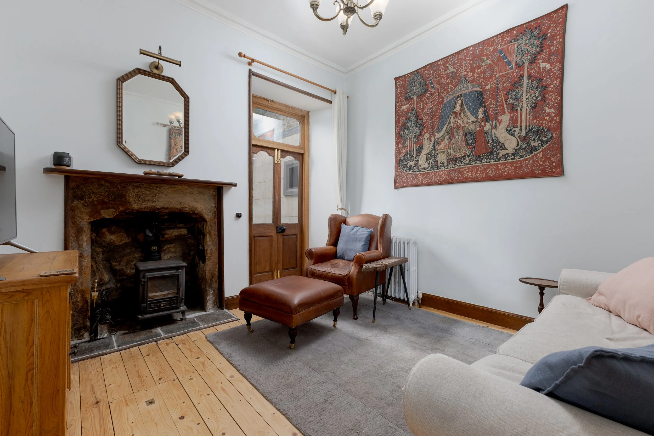 A cozy living room interior with a stone fireplace, a wooden mantle, and a wood-burning stove. There is a leather armchair with a matching footrest, a gray rug, a large tapestry on the wall, and a radiator. A door with glass panels and white curtains