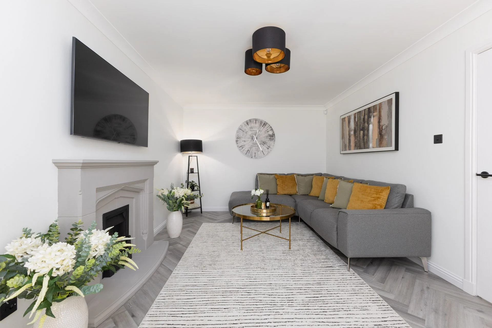 Living room with gray sofa, gold-trimmed glass coffee table, white and black decor, wall art, large TV, black ceiling light, and white walls.