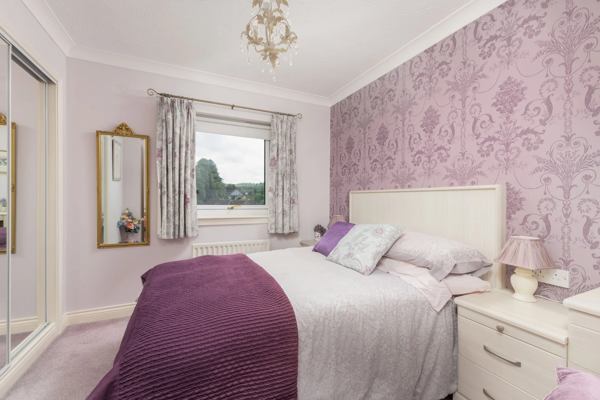 A bedroom with lavender and white floral wallpaper on one wall, a white headboard, two white side tables with lavender lamps, a window with floral curtains, and a purple bedspread and pillows.