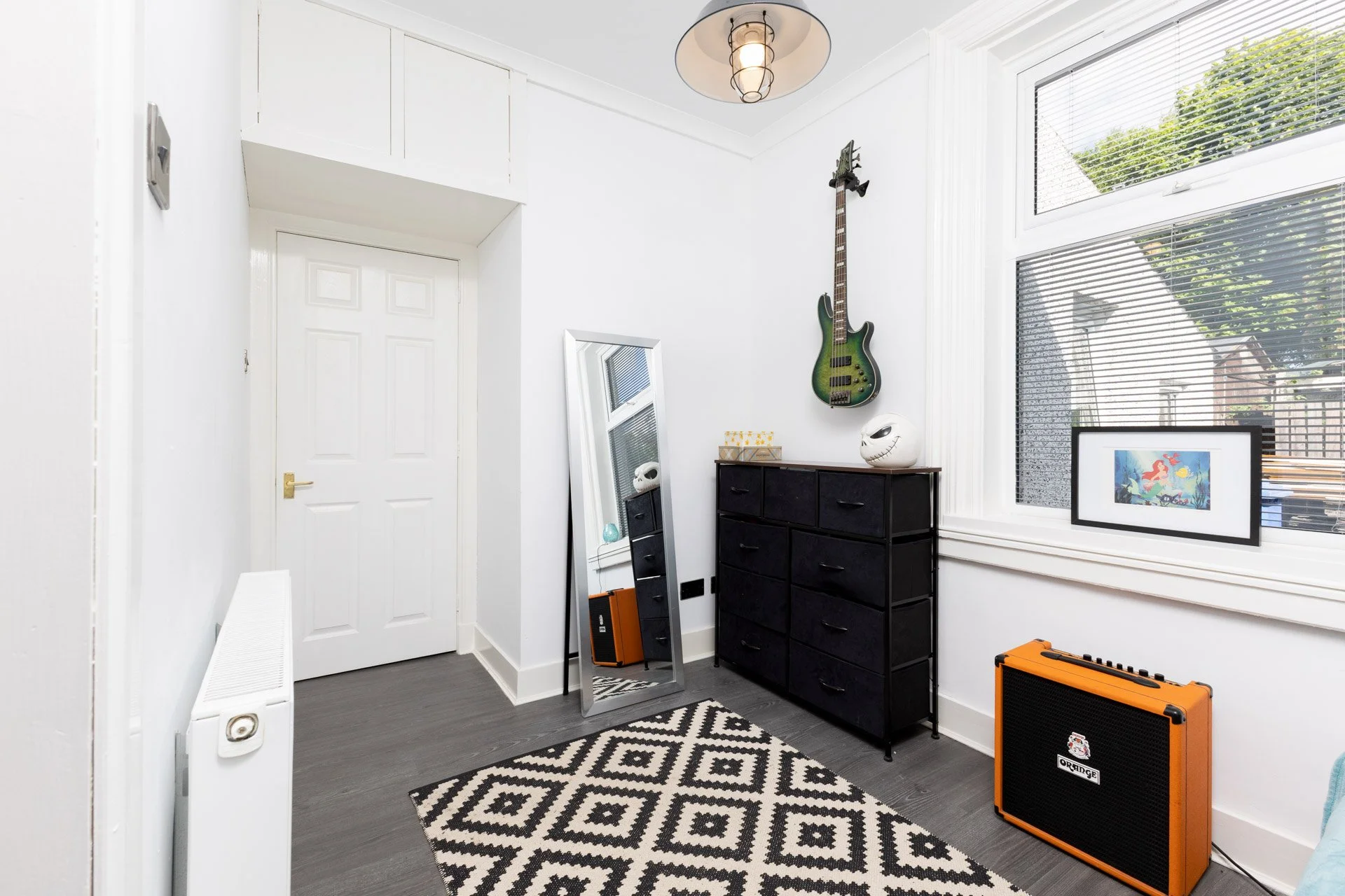 Interior of a room with white walls, a white door, a black dresser, a standing mirror, an electric guitar mounted on the wall, a framed artwork, and musical equipment including an orange amplifier and a black speaker cabinet. A black and white patter
