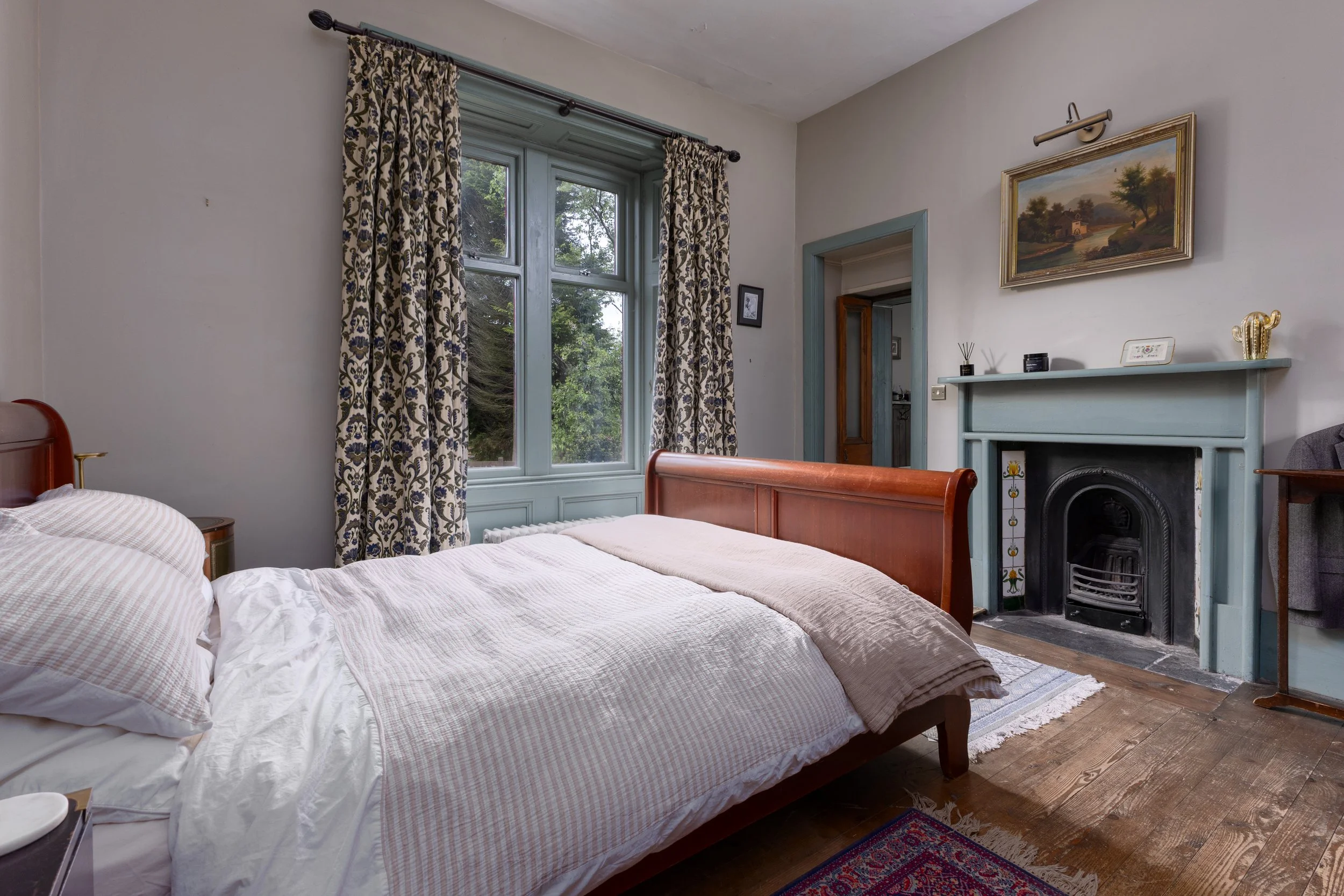 A cozy bedroom, a window with patterned curtains, and a fireplace with a beautiful fire surround. The room has wooden floors and a small rug, with artwork hanging on the wall.