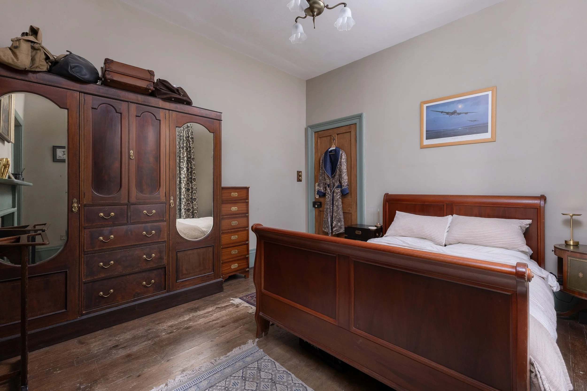 A domestic bedroom with a wooden bed frame, white bedding, and a bedside table with a brass lamp. There is a large wooden wardrobe with bags stored on top, a wooden dresser, and a small mirror. A door has a coat or robe hanging on it. The walls are l