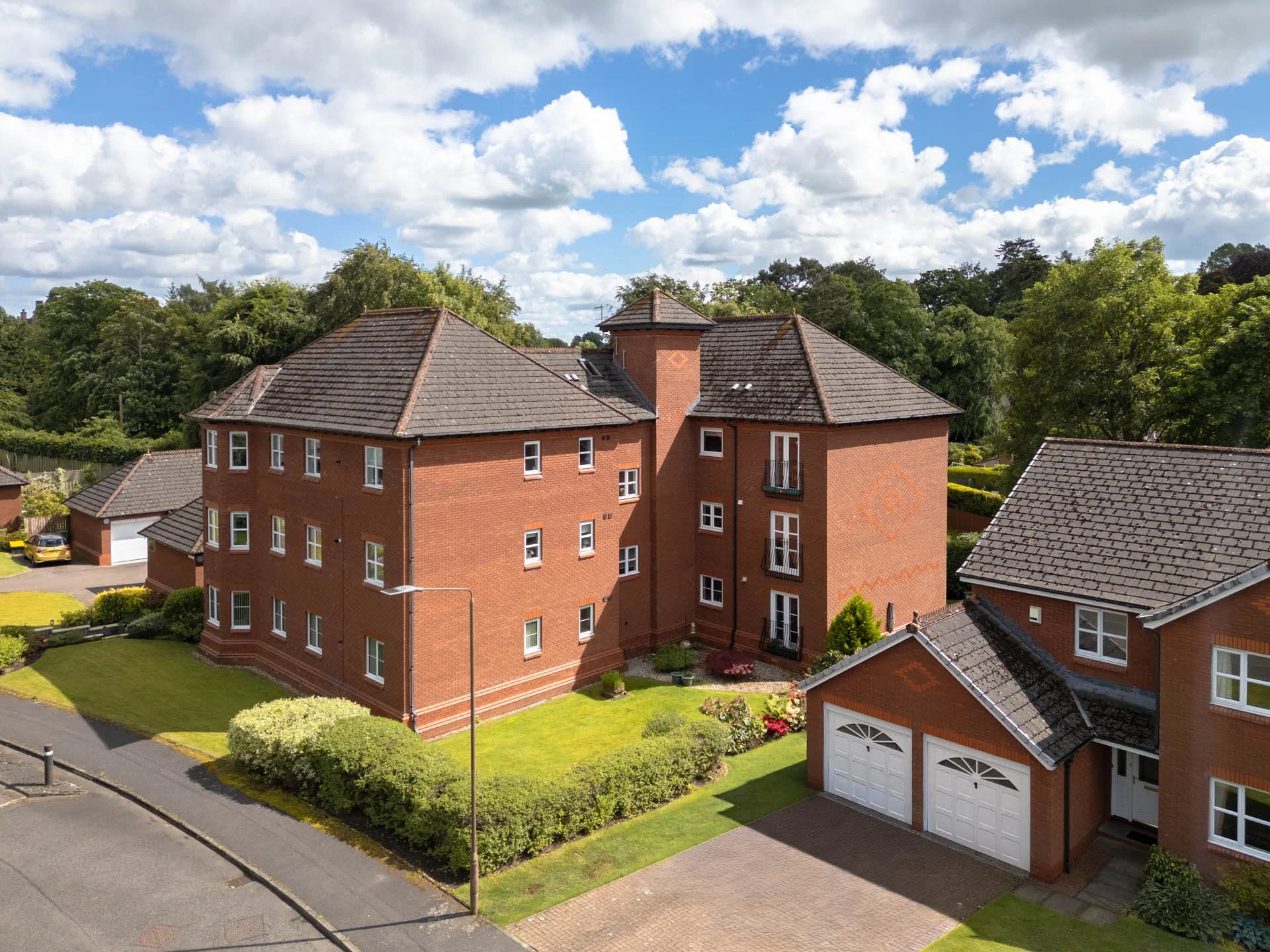 View of a modern red brick residential building with multiple windows and front gardens, surrounded by lush green trees under a cloudy sky.