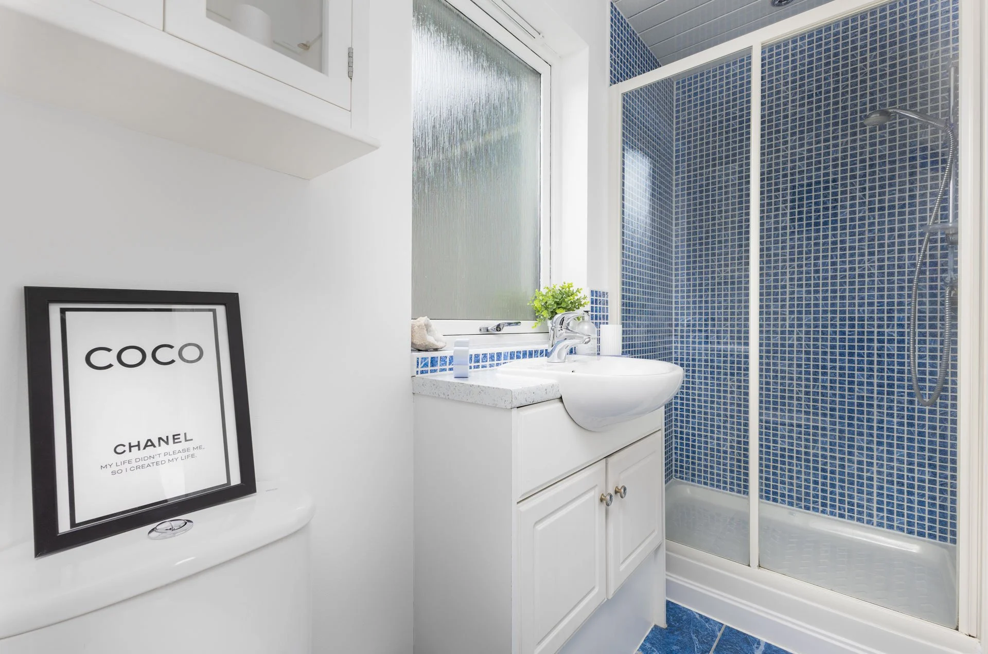 A small bathroom with a white vanity, blue mosaic tiled shower enclosure, and a framed Chanel print on the wall.