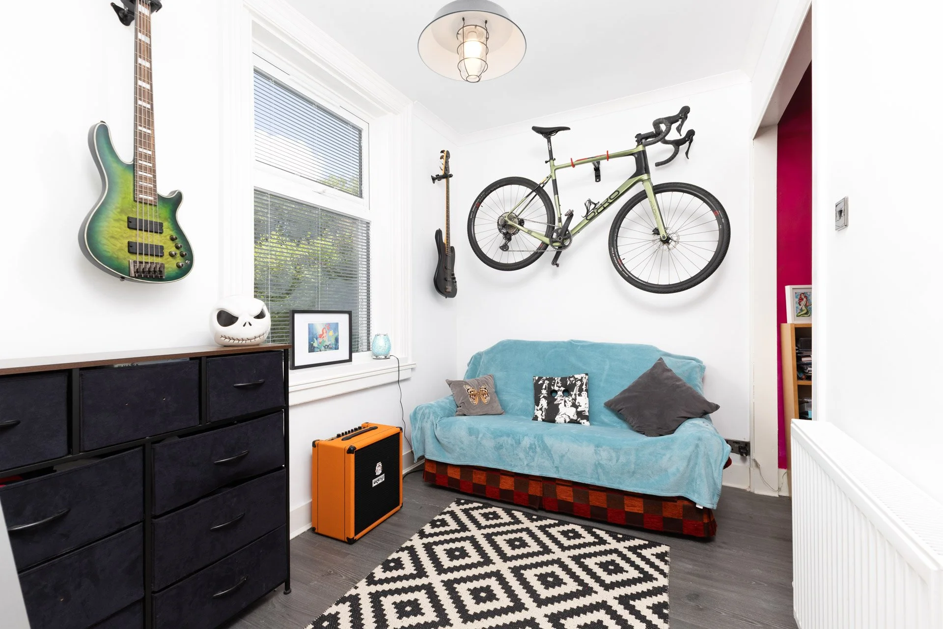 A living room corner with a blue sofa, a black dresser, and a window. Wall-mounted on the wall are a green bass guitar and a black bicycle. There is a small framed picture on the window sill, a skull-shaped lamp, and an orange amplifier on the floor.
