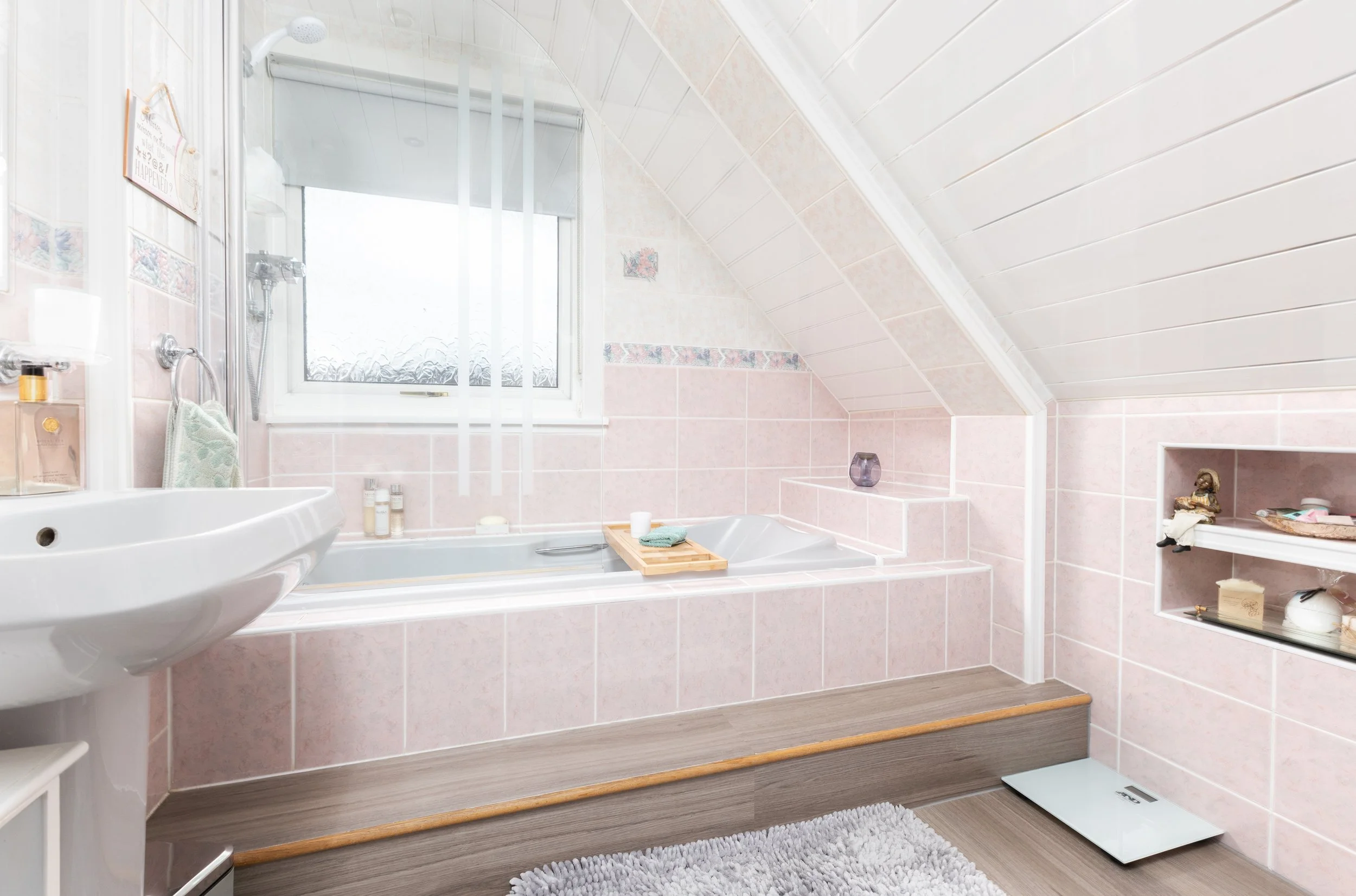 A bathroom with a bathtub, a sink, a window with frosted glass, and built-in shelves. The wall and floor tiles are pink and neutral tones. There are toiletries, a scale, and decorative items placed around.