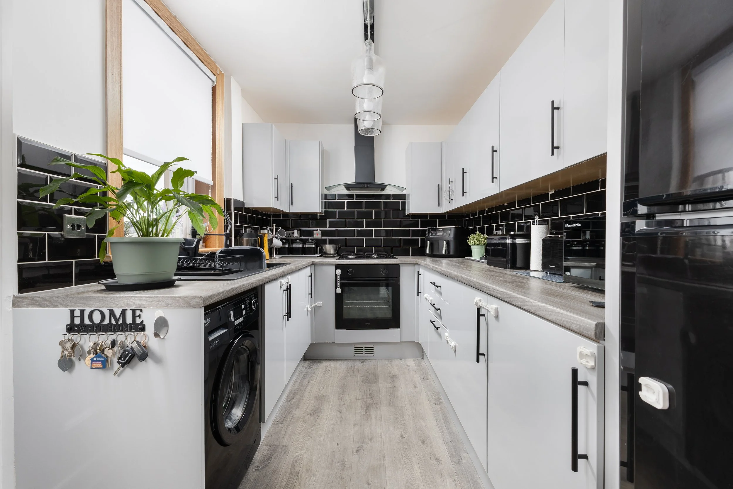 A modern U-shaped kitchen with white cabinets, black hardware, black backsplash tiles, stainless steel appliances, a small potted plant, a window with a blind, and a wooden countertop. There is a washing machine, a stove, and a microwave visible.