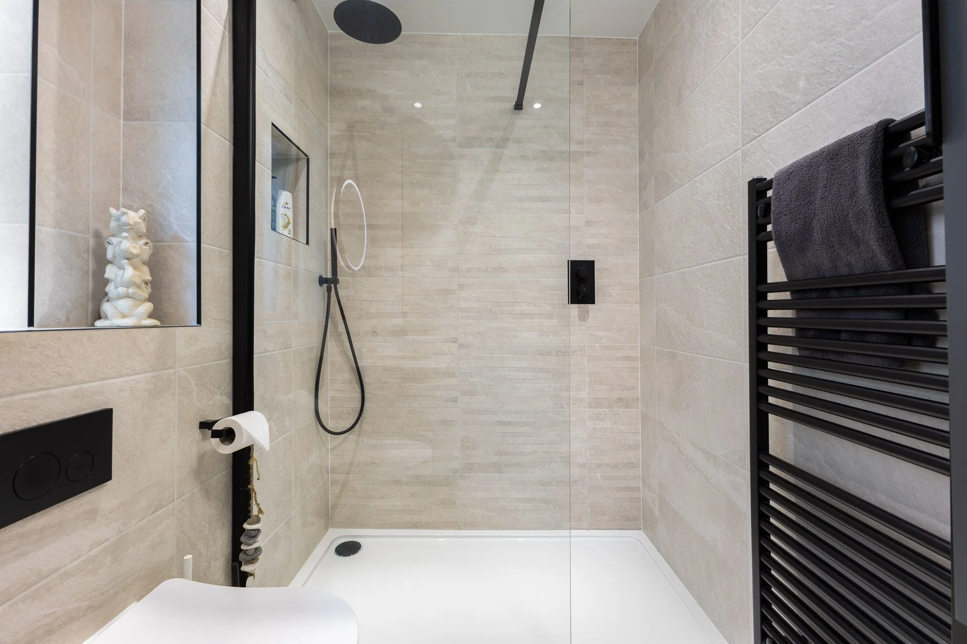 Modern minimalist shower with beige tile walls, black fixtures, rain showerhead, handheld shower, built-in niche with toiletries, black towel radiator with dark towel, and a white shower tray.