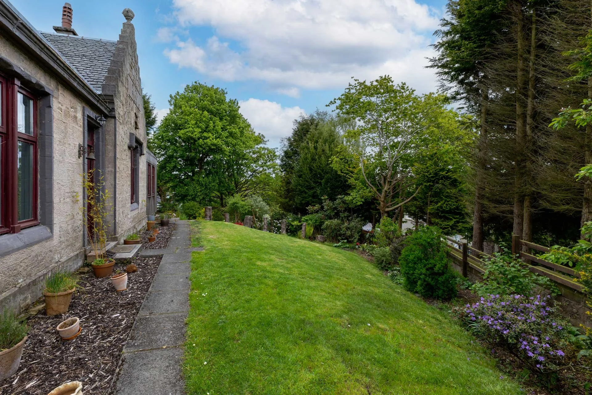 A backyard garden with a stone house on the left, a pathway with potted plants along it, a lush green lawn, and a variety of trees and shrubs on the right under a partly cloudy sky.