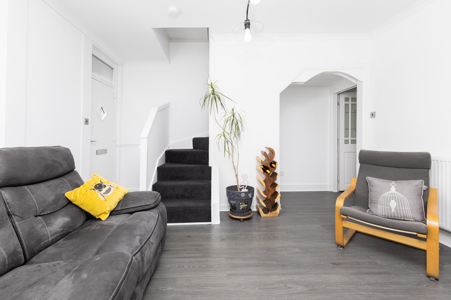 Interior of a living room with a gray couch on the left with a yellow pillow, a gray armchair with a decorative pillow on the right, a small black table with stickers and a pink cat figurine, a wooden wine rack, a tall potted plant, a staircase with 