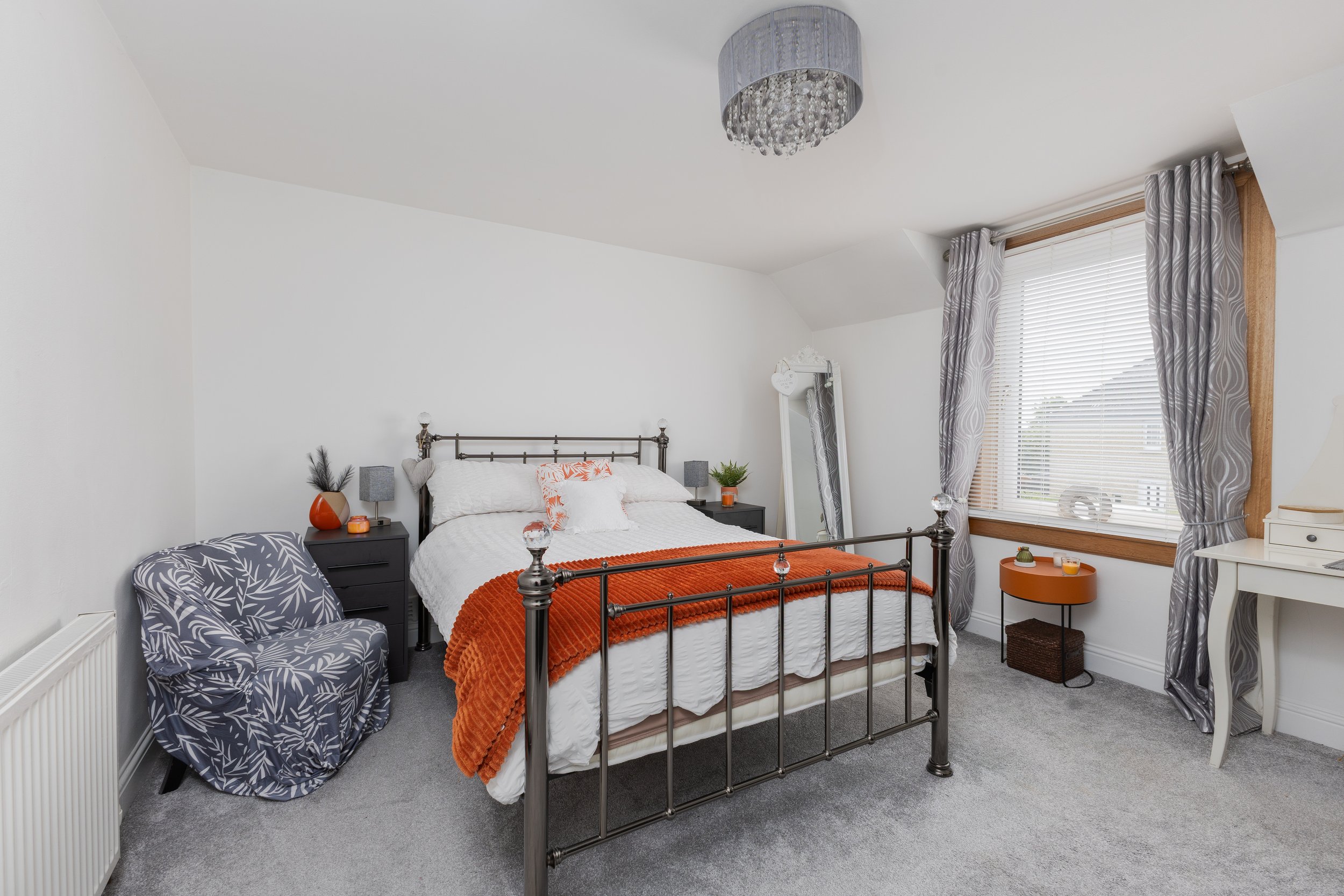 A cozy bedroom with a black metal bed frame, white bedding, and orange throw blanket. There's a plaid armchair with a leaf pattern, two black nightstands with lamps, and a large window with gray and white patterned curtains. A chandelier hangs from t
