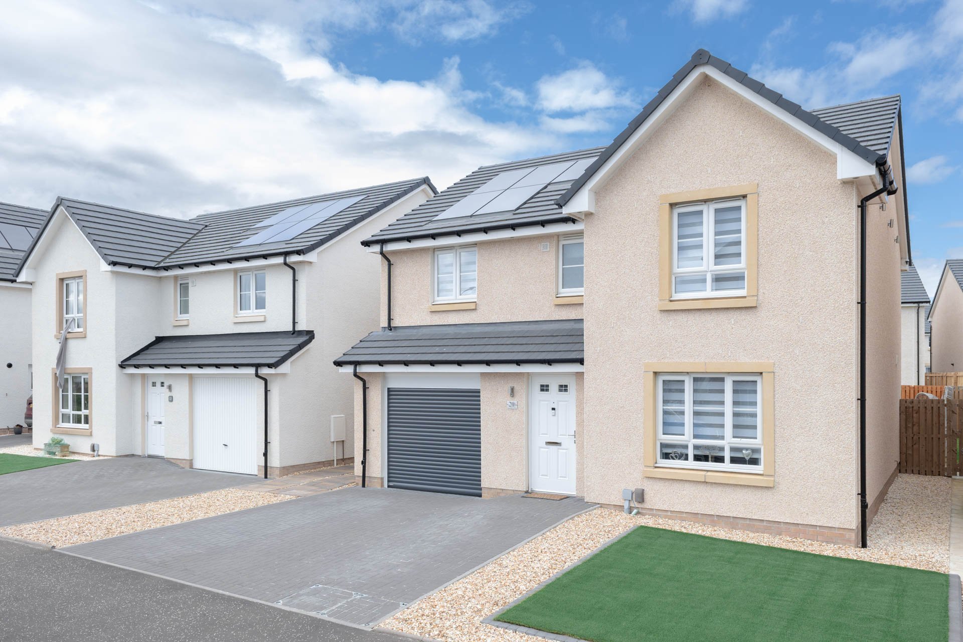 Adjacent modern suburban homes with solar panels on the roofs, driveways, and well-maintained lawns under a partly cloudy sky.