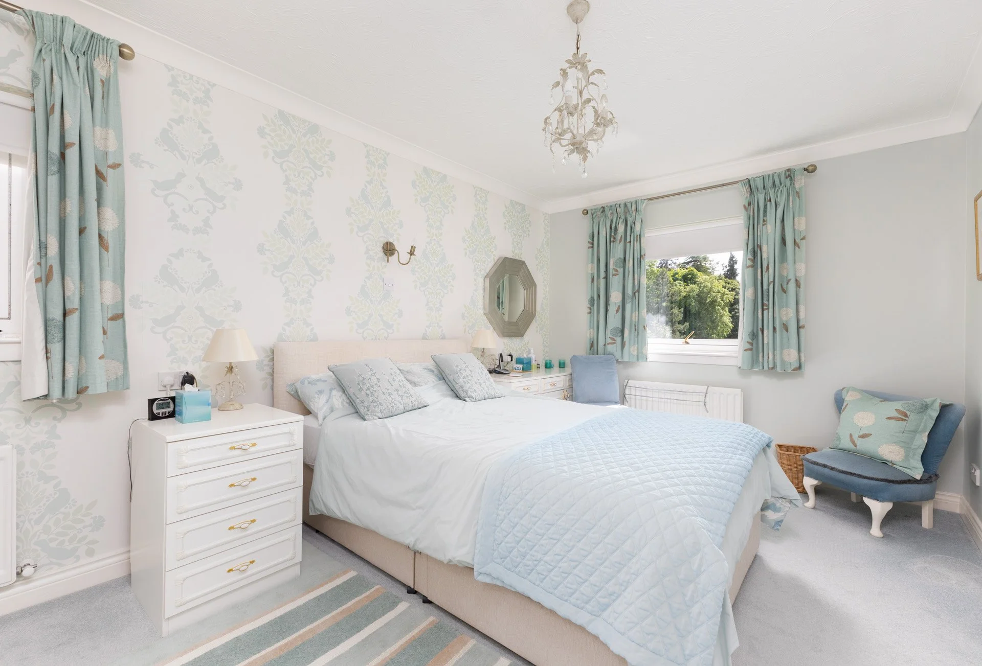 A bedroom with a white bed, bedside table with a lamp, and light blue and white decor, featuring curtains with a bird and leaf pattern, a chandelier, a mirror on the wall, a window showing green trees outside, and a chair with matching throw pillows.