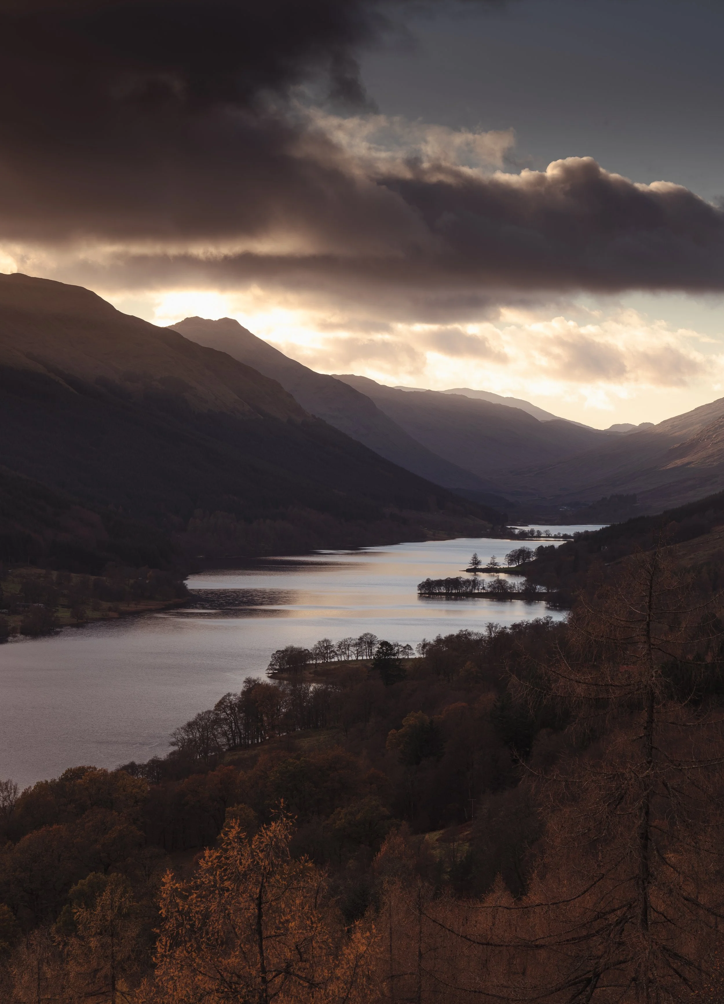 A scenic landscape of a river flowing through a valley surrounded by mountains with a partly cloudy sky at sunset or sunrise.
