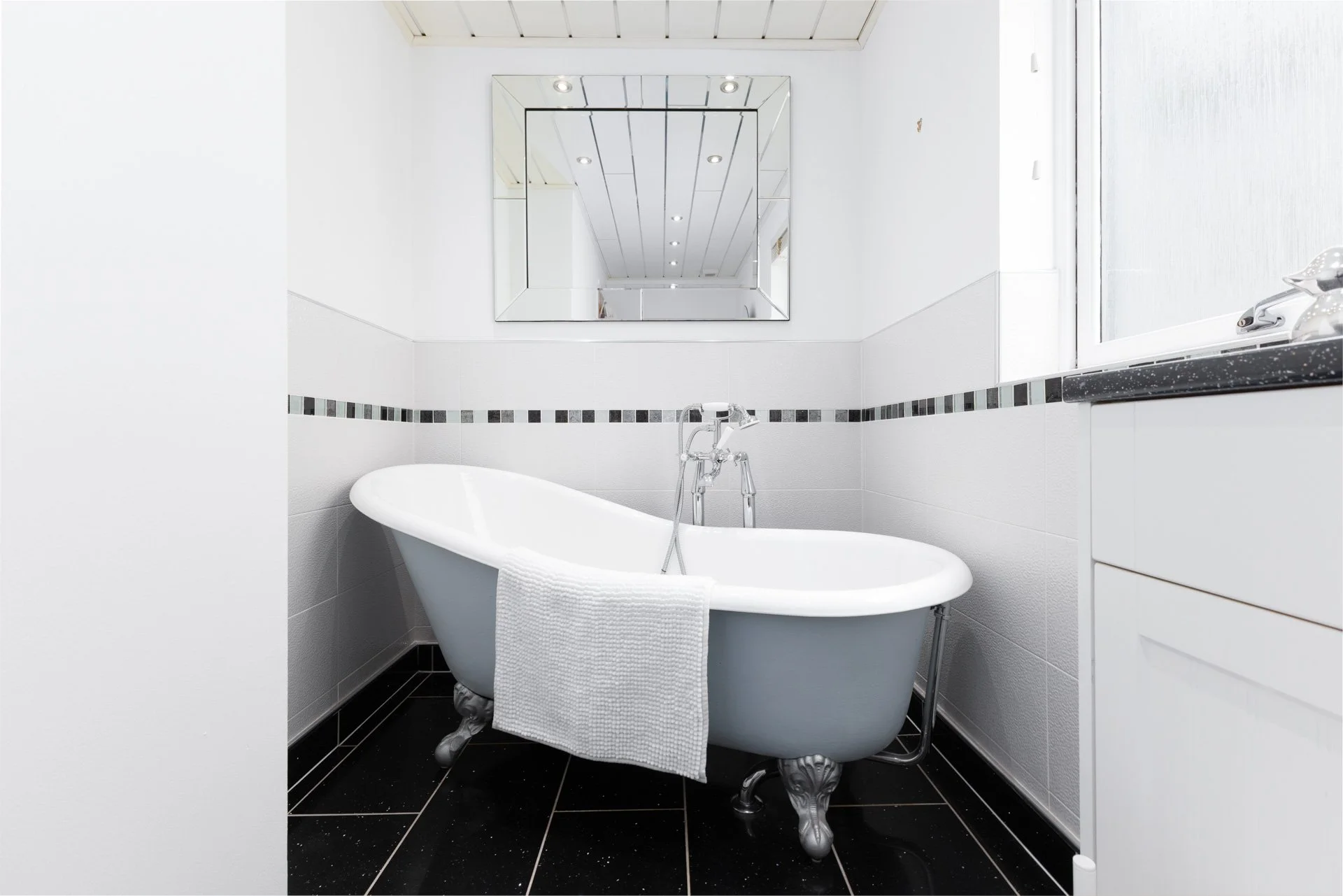 A modern bathroom with a white bathtub and decorative clawfoot feet, a white towel hanging over the side, a large wall mirror, a window with frosted glass, and black and white tiled walls and floors.