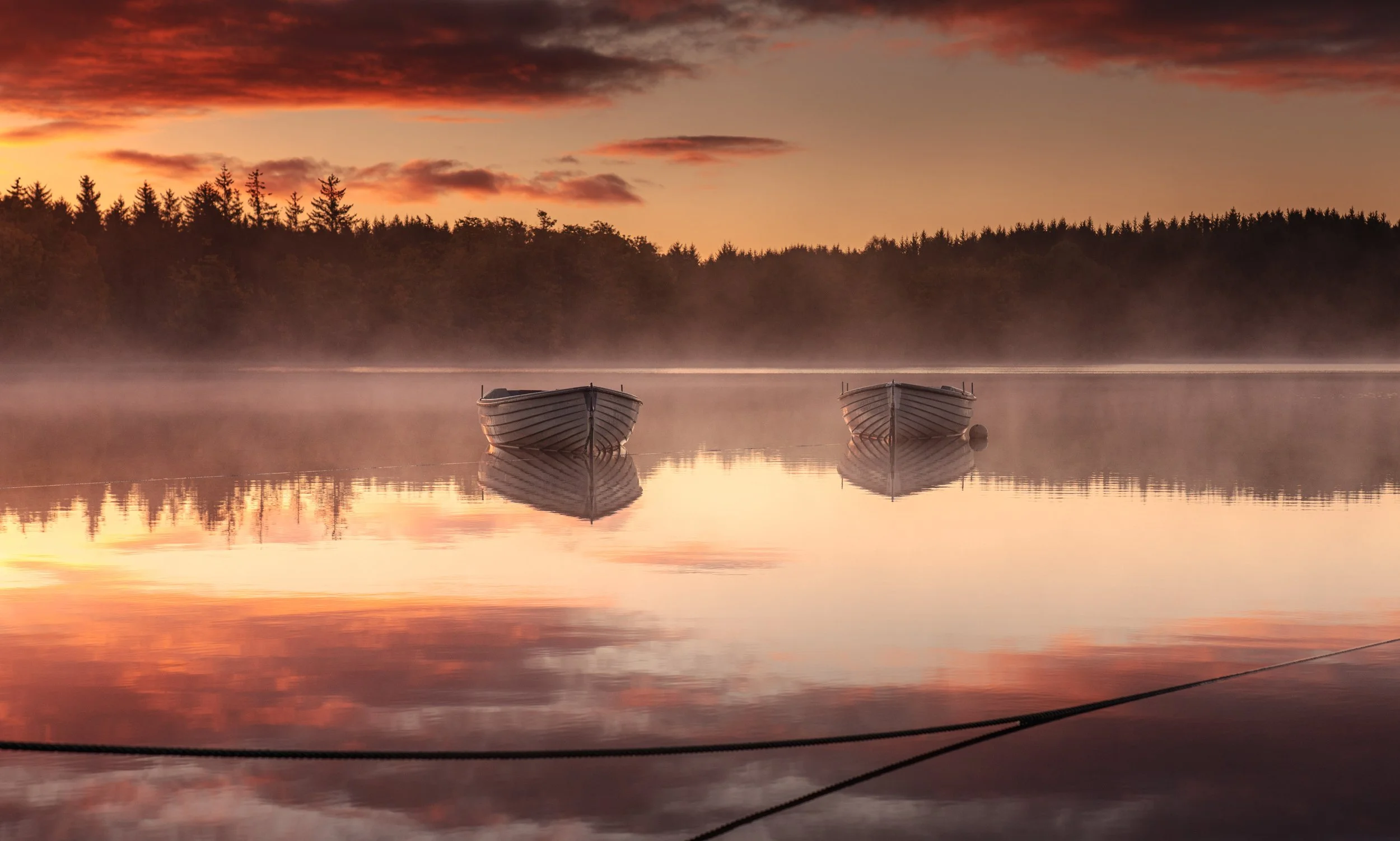 Two boats floating on a calm lake during sunset with a misty atmosphere and a forested shoreline in the background.