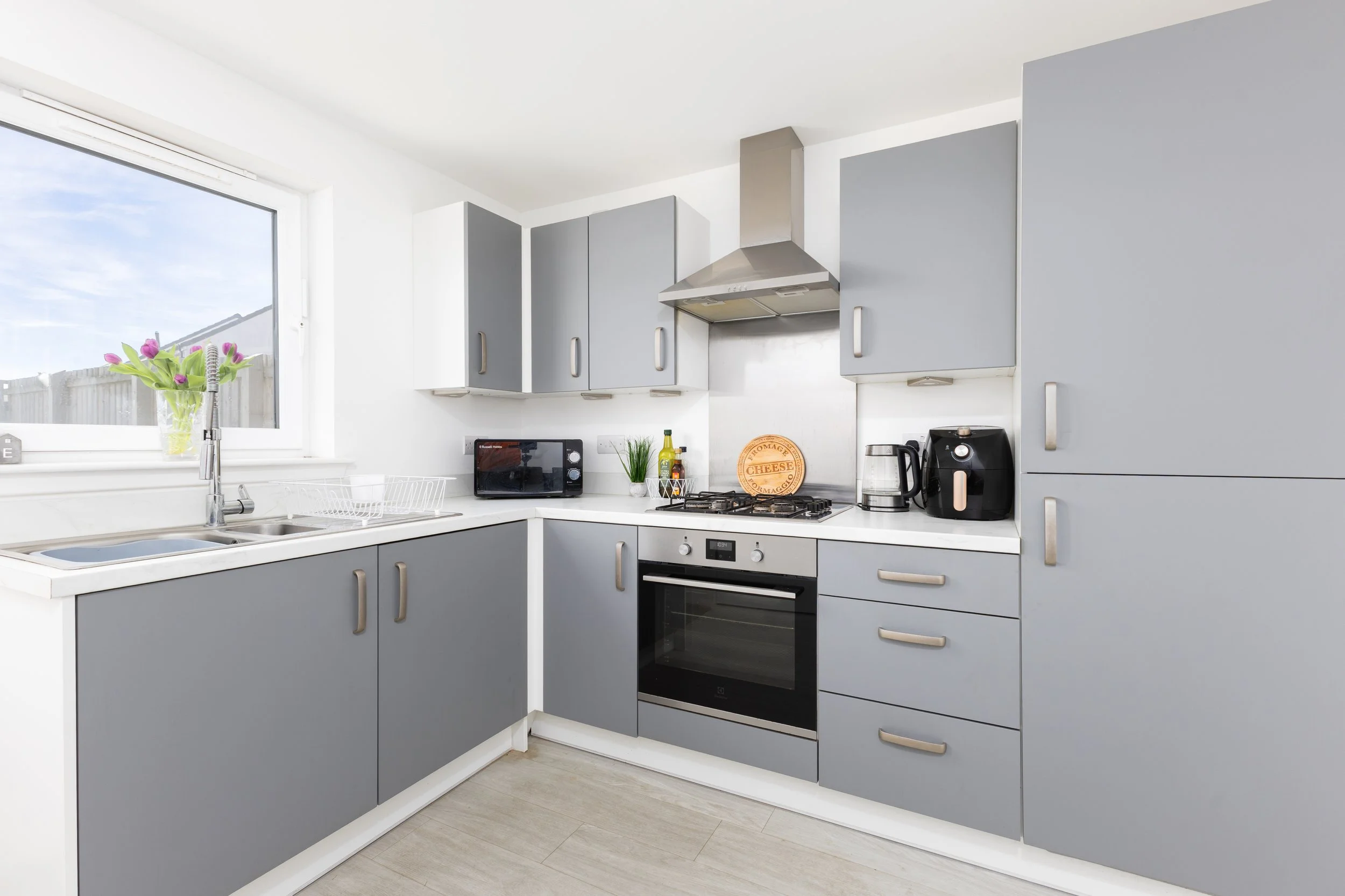 Modern kitchen with gray cabinets, a window with a vase of pink tulips, a countertop with a microwave, kettle, and condiments, and stainless steel appliances including a stove and range hood.