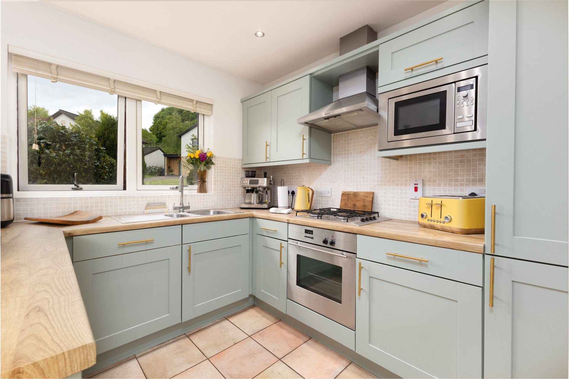 Kitchen with pastel green cabinets, wooden countertops, a window overlooking greenery, and various appliances including a microwave, toaster, kettle, coffee maker, and oven.