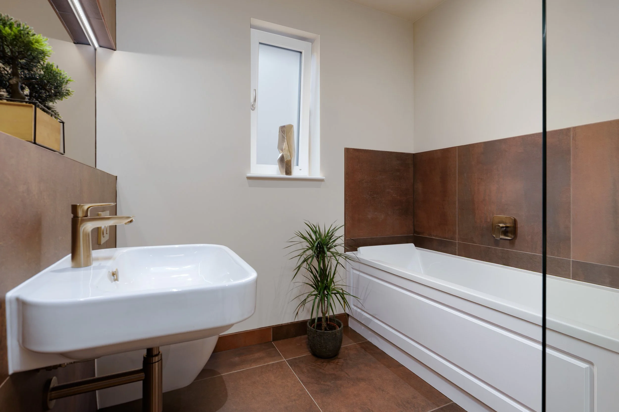 A modern bathroom features a white bathtub, a white sink with a gold faucet, and a small green plant in a black pot. The walls have white upper sections and brown tiles lower sections, with a frosted window above the bathtub. There is a decorative sc