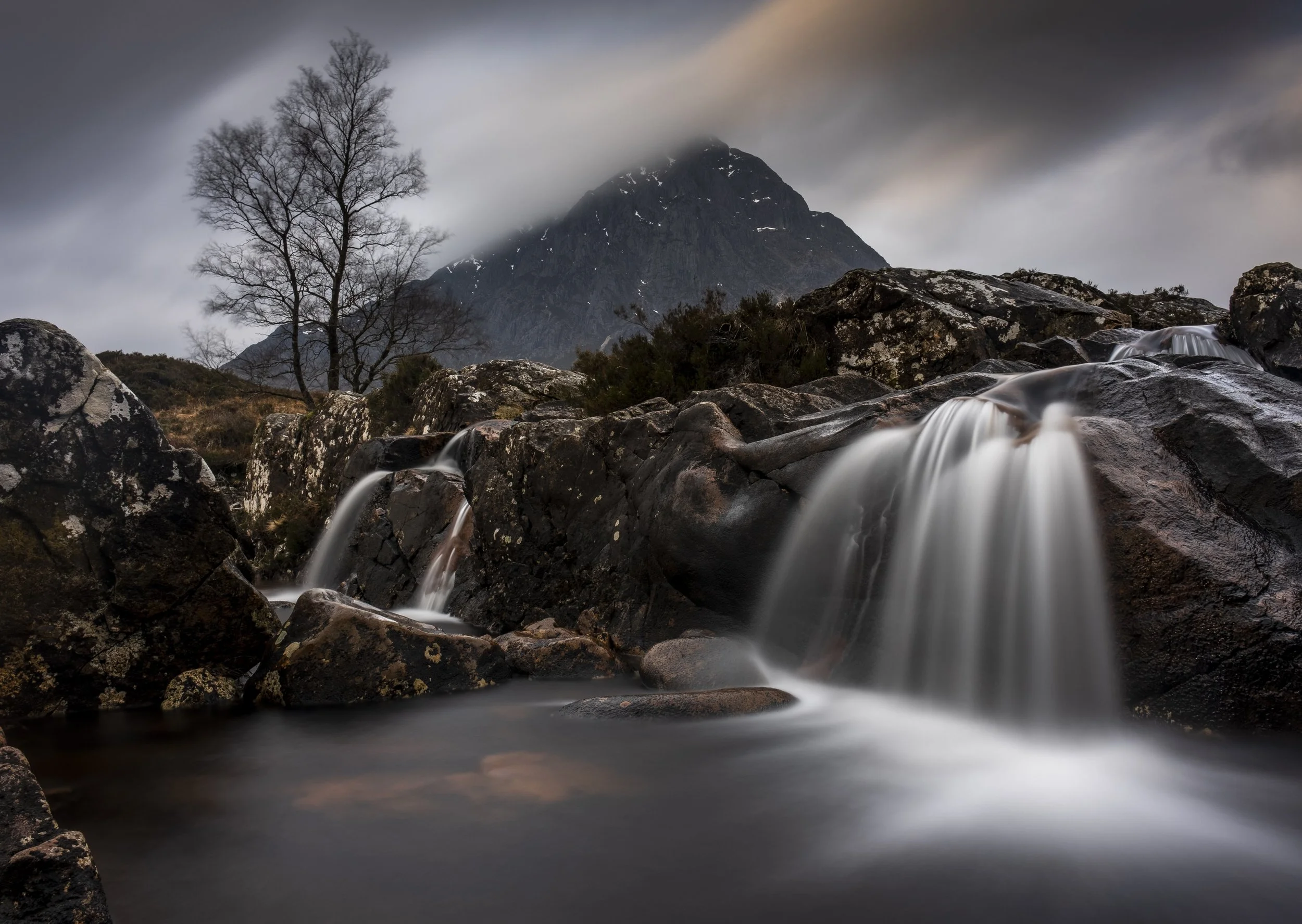 A mountain with snow patches partially obscured by clouds, a leafless tree, and a small waterfall flowing over rocks in a rugged landscape under a cloudy sky.