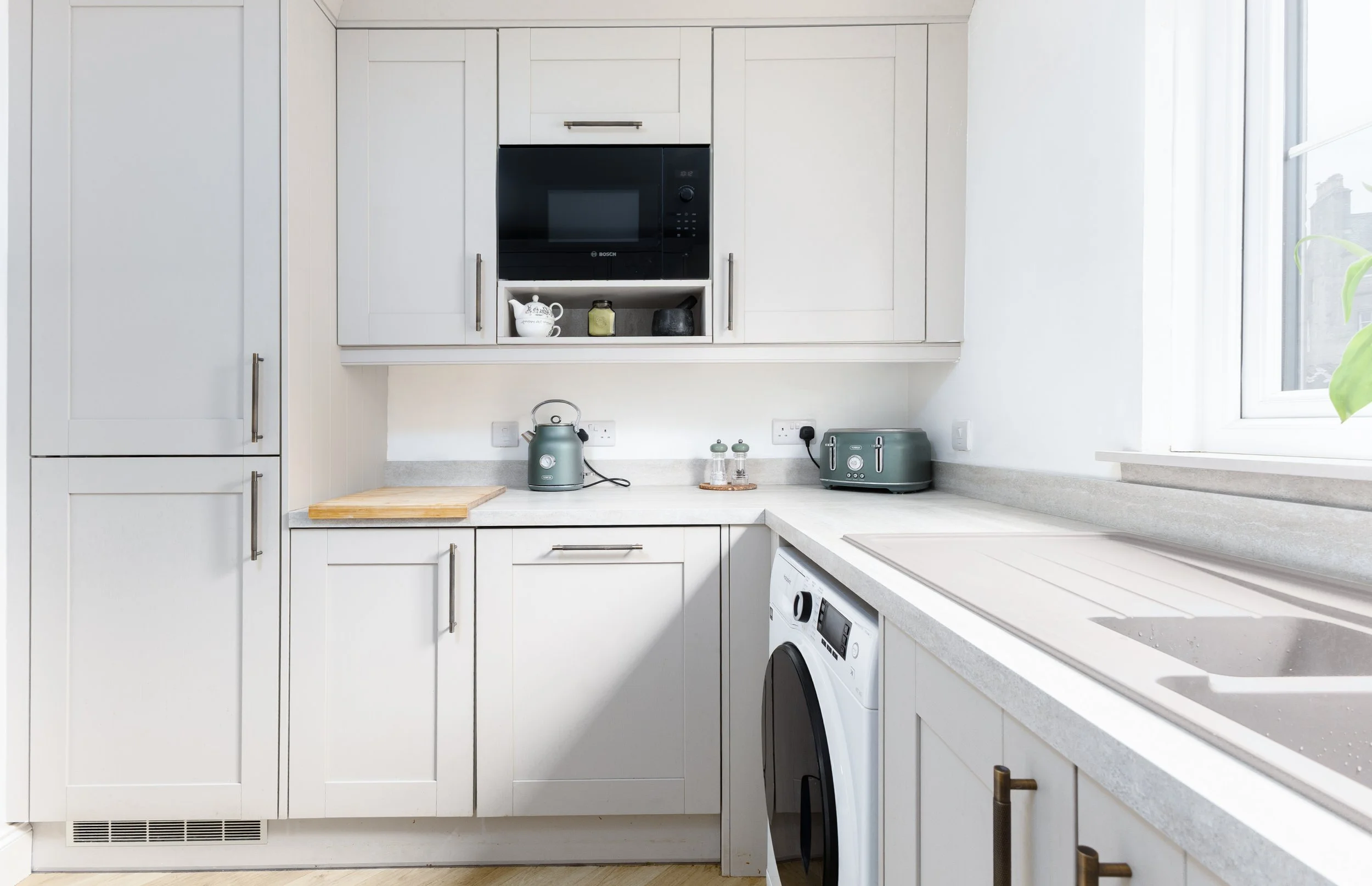 A modern kitchen with white cabinets, a microwave oven, a washing machine, and small appliances on the countertop. There is a window on the right side with natural light, and a small shelf with decorative items above the countertop.