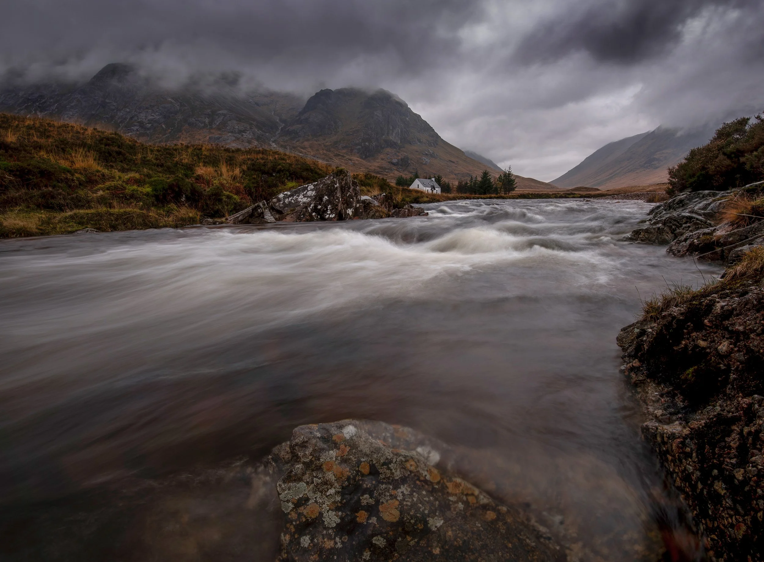 A river flowing rapidly through a mountainous landscape with cloudy, overcast skies and a small white house with a dark roof on the riverbank.
