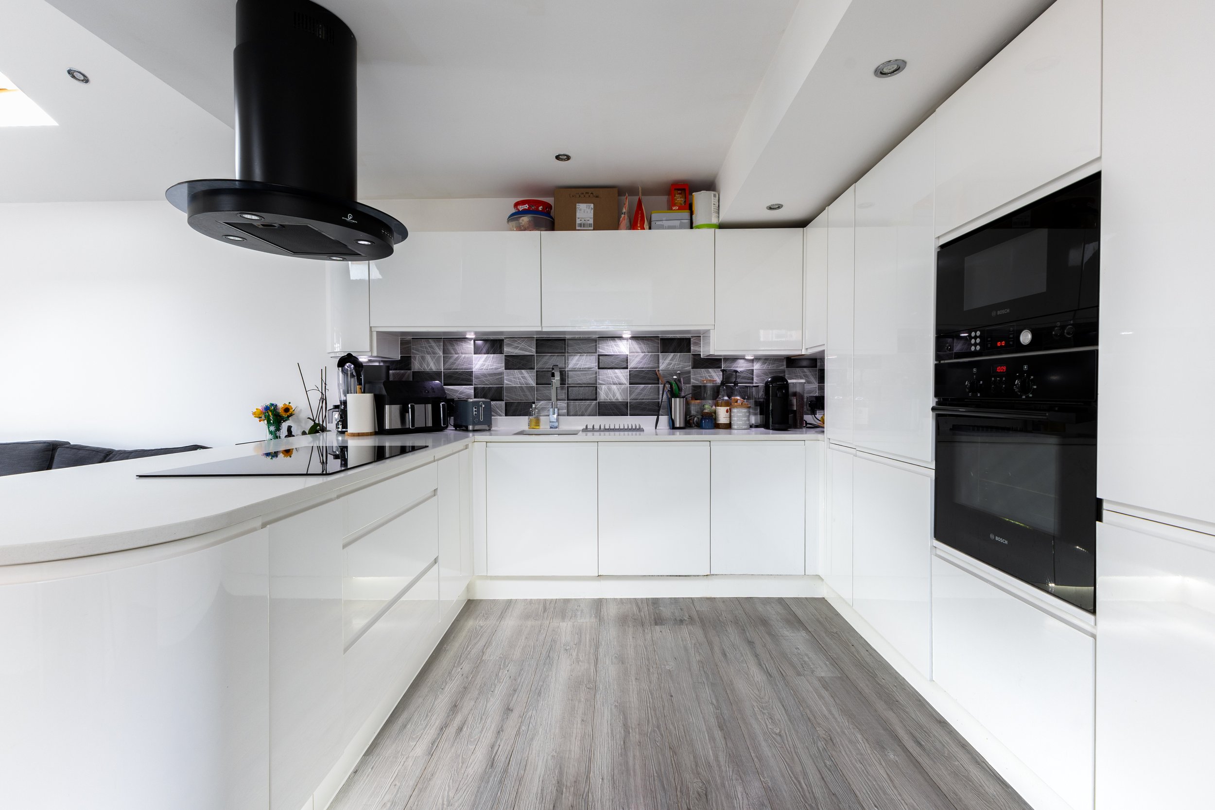 Modern white kitchen with black appliances, gray tile backsplash, and gray wood flooring.