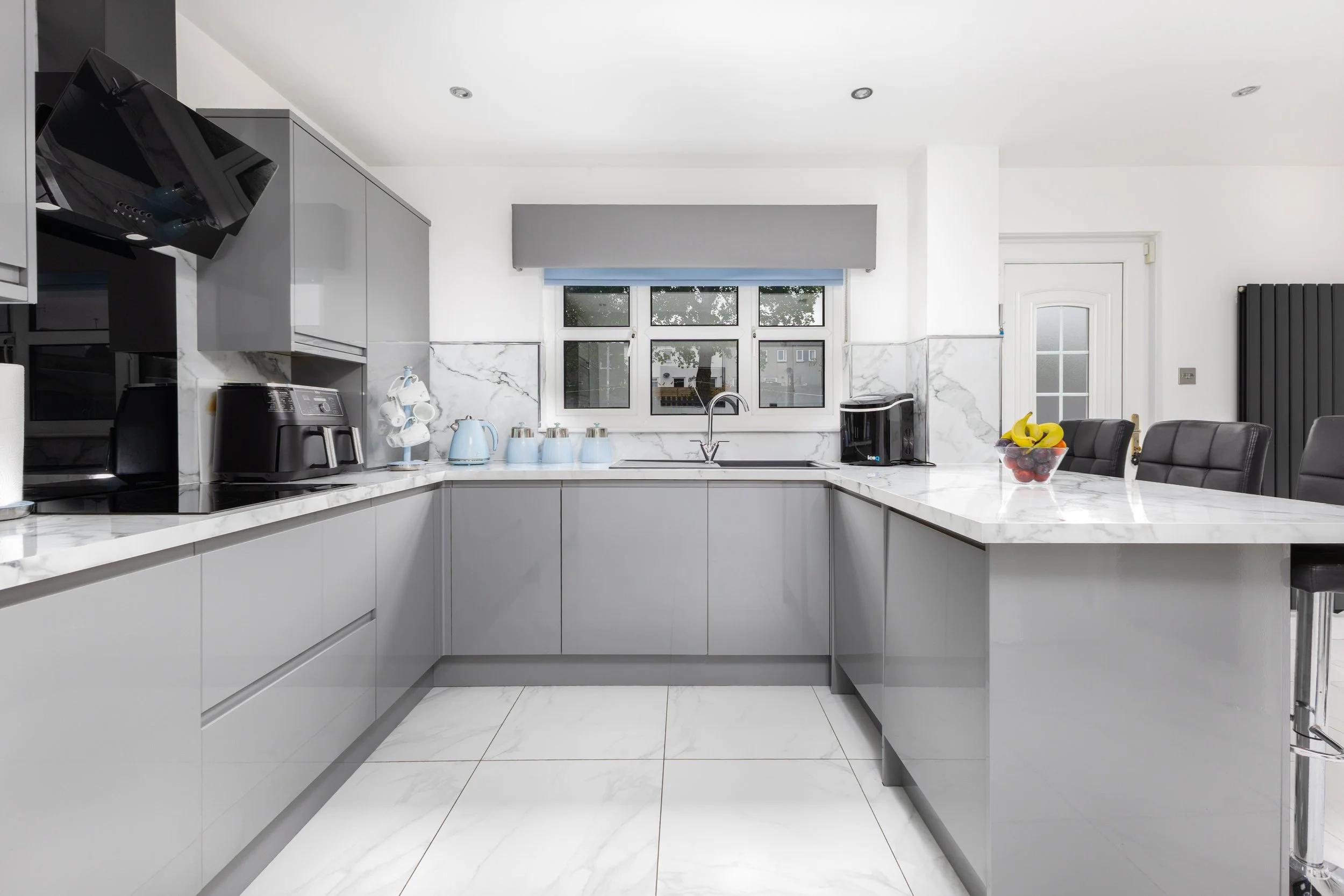 Modern kitchen with white marble countertops and backsplash, gray cabinetry, a window with a gray blind, and various small appliances including a coffee maker, electric kettle, and a fruit bowl containing bananas, strawberries, and grapes.