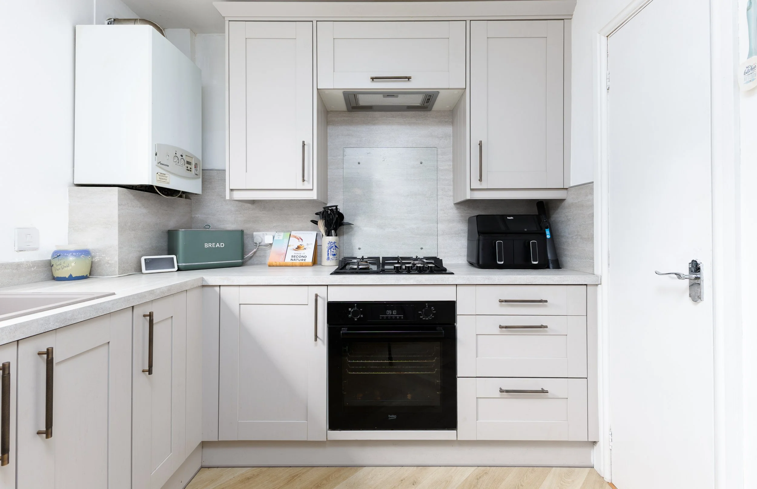 A white kitchen with upper and lower cabinets, a black oven, a stovetop, a range hood, a toaster, a small speaker, a cookbook, utensils in a holder, and a white door on the right side.