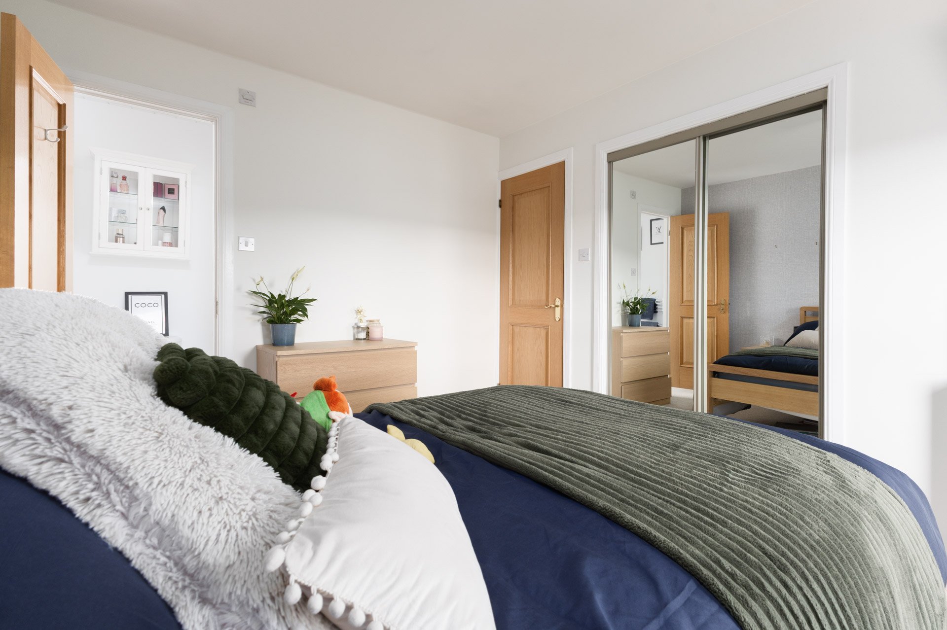 A bedroom with a bed covered in a dark green blanket and decorated with plush toys, including a black fluffy toy and a small orange and green toy. There is a white furry pillow with pom-pom trim. The room has white walls, a wooden door, and a mirrore