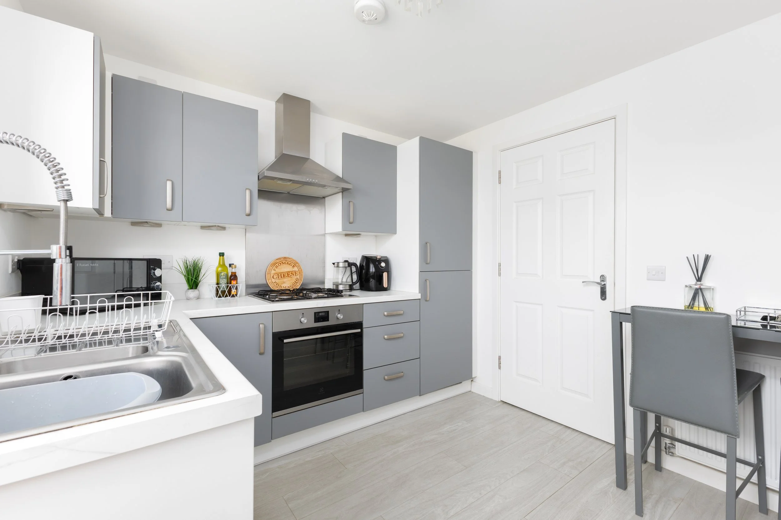 Modern kitchen with gray cabinets, stainless steel range hood, black oven, and small dining area with gray chairs.