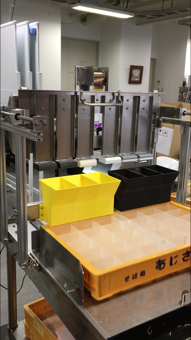 A metal confectionery machine in a workshop setting, with yellow and black containers on a wooden tray in front of it, and a large tray with multiple compartments on the table.