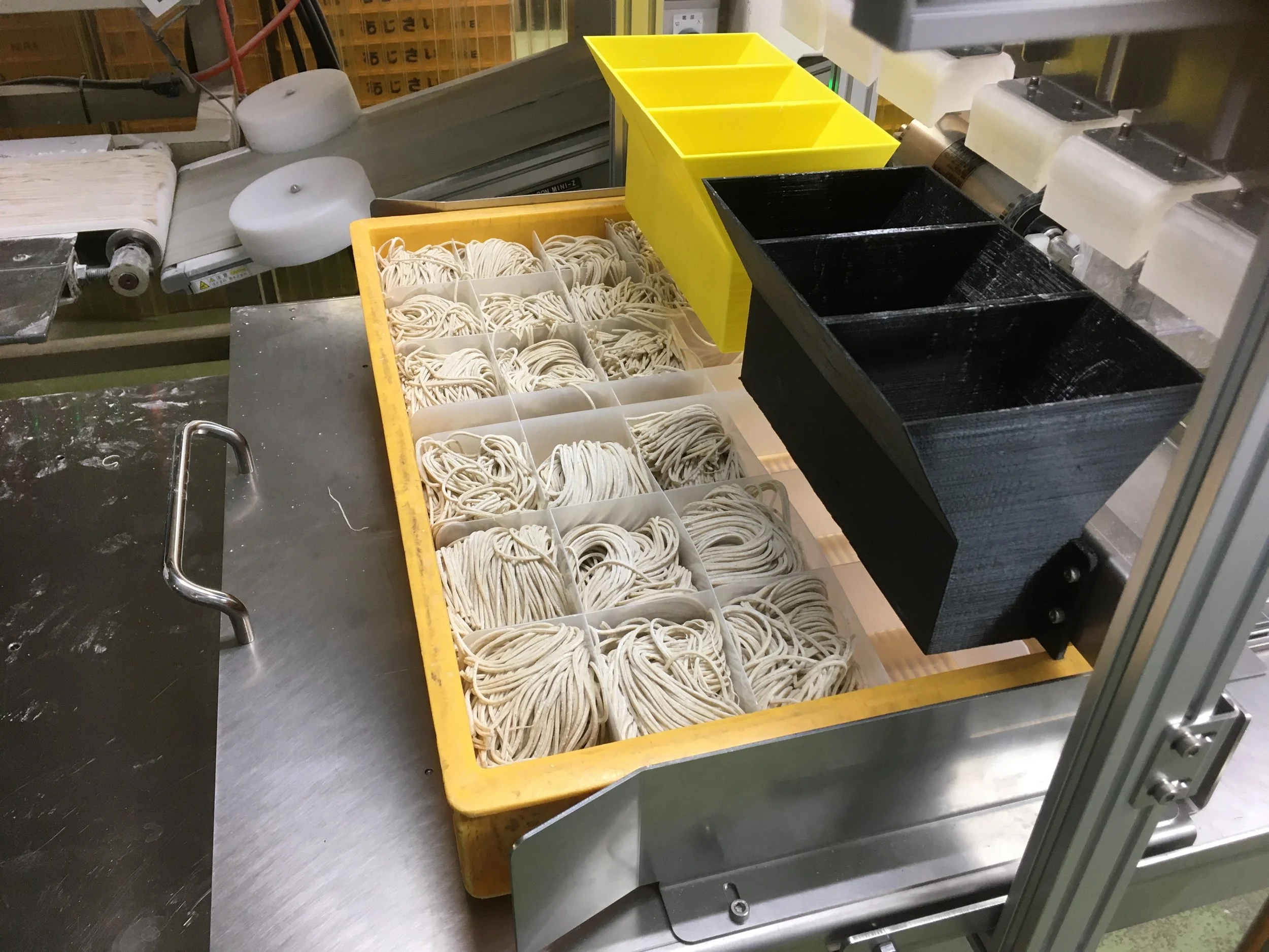 A commercial noodle-making machine with yellow and black bins filled with freshly made noodles.