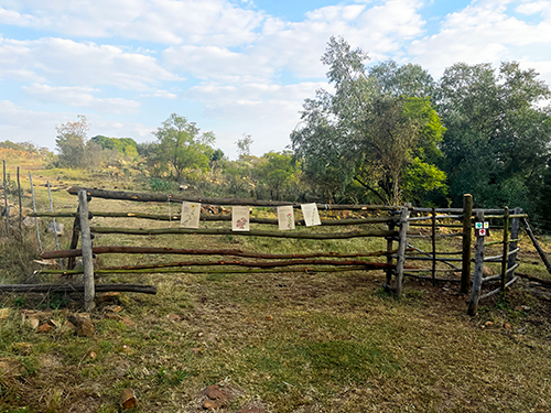 Rustic wooden gate across a dirt path with signs hanging from the top rail, surrounded by trees and grassy land under a partly cloudy sky.