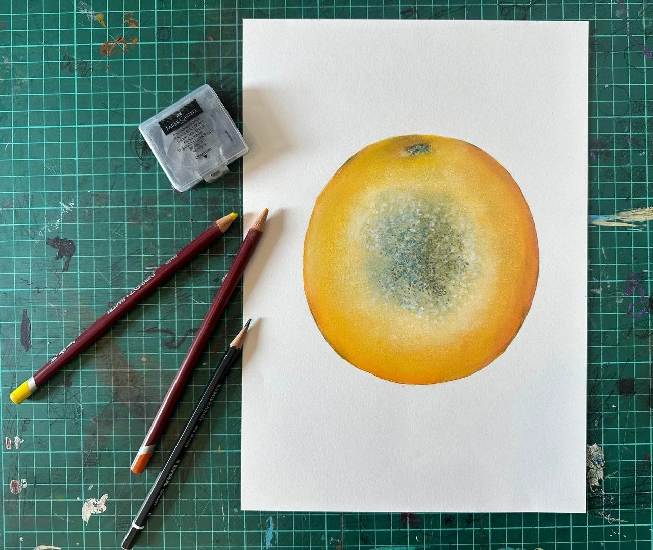 Illustration of an orange, with the image placed on white paper on a green cutting mat, along with soft pastel pencils.