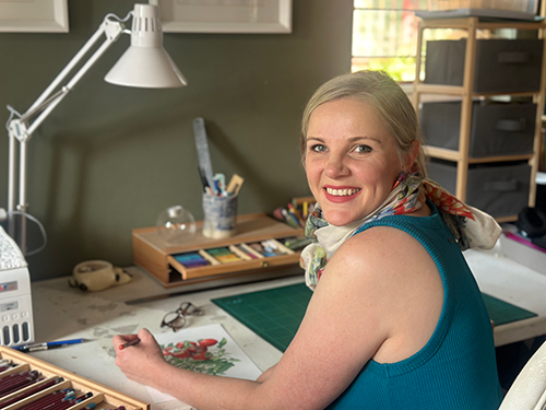 A woman with blonde hair smiling while working on an illustration in her studio.